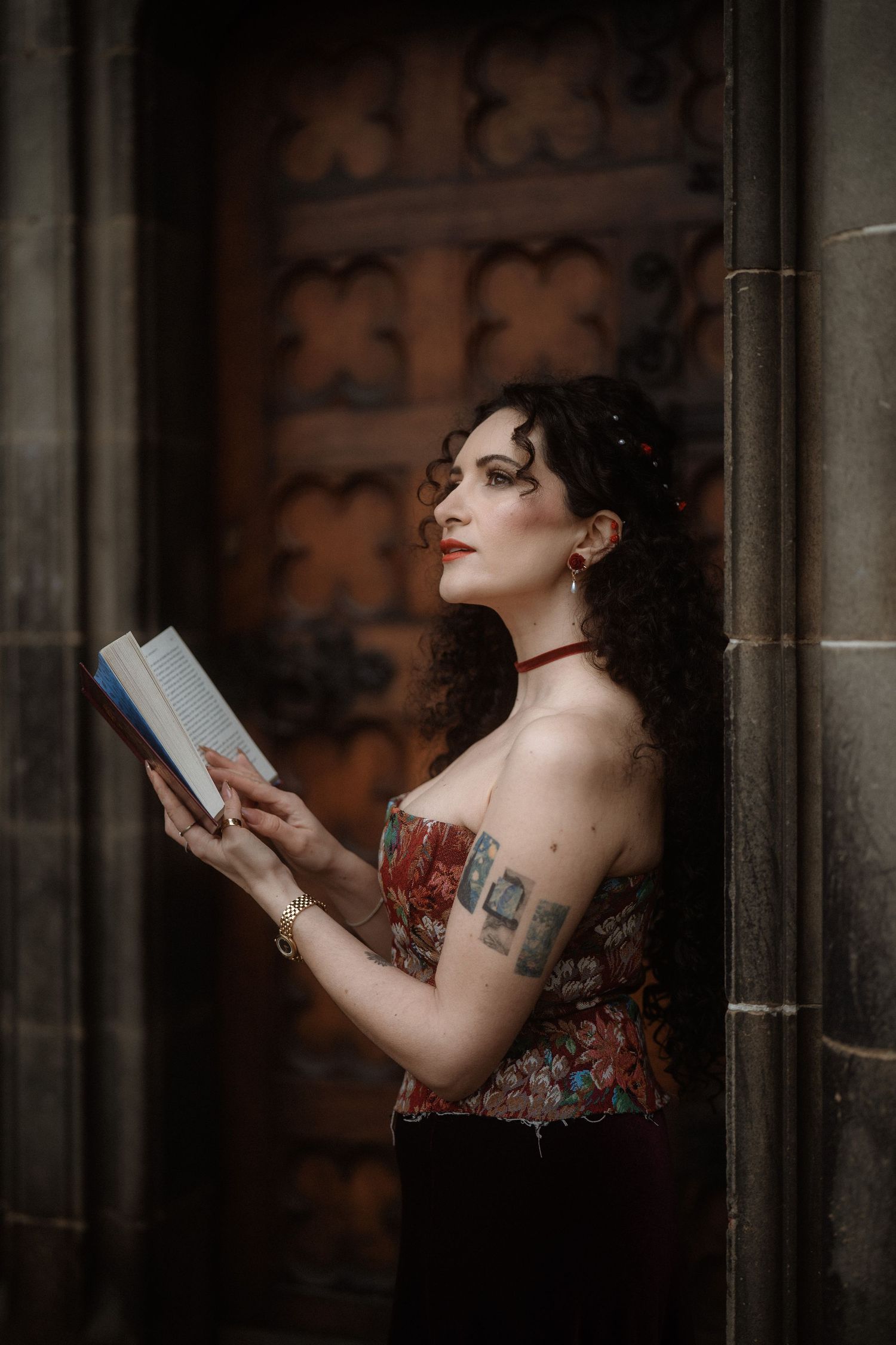 Woman with curly dark hair, red lips, and tattoos holds a book while leaning against a stone wall near an ornate door.