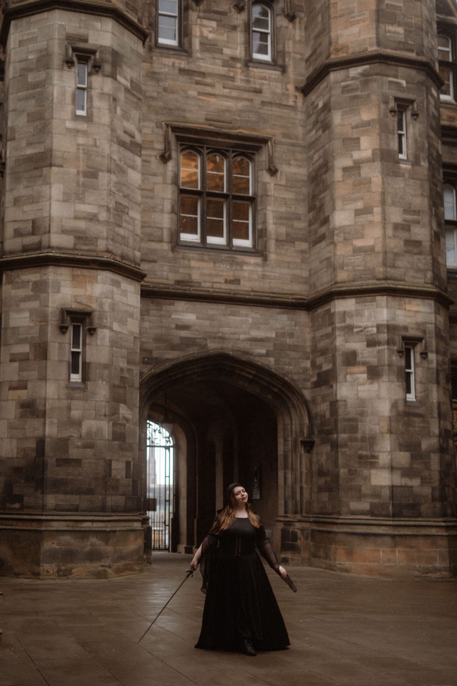 A figure in a dramatic black gown stands beneath a Gothic stone archway at a historic castle or university building.