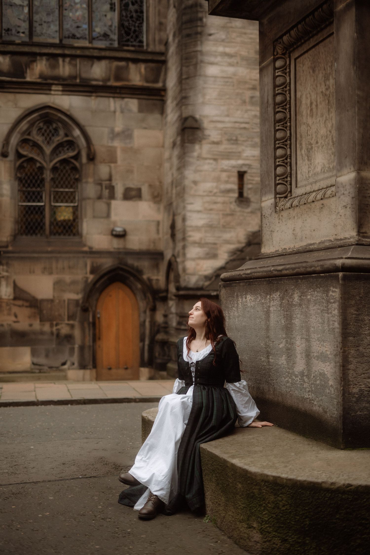 A figure in a white dress and dark top sits on stone steps against a historic church wall with Gothic architecture.