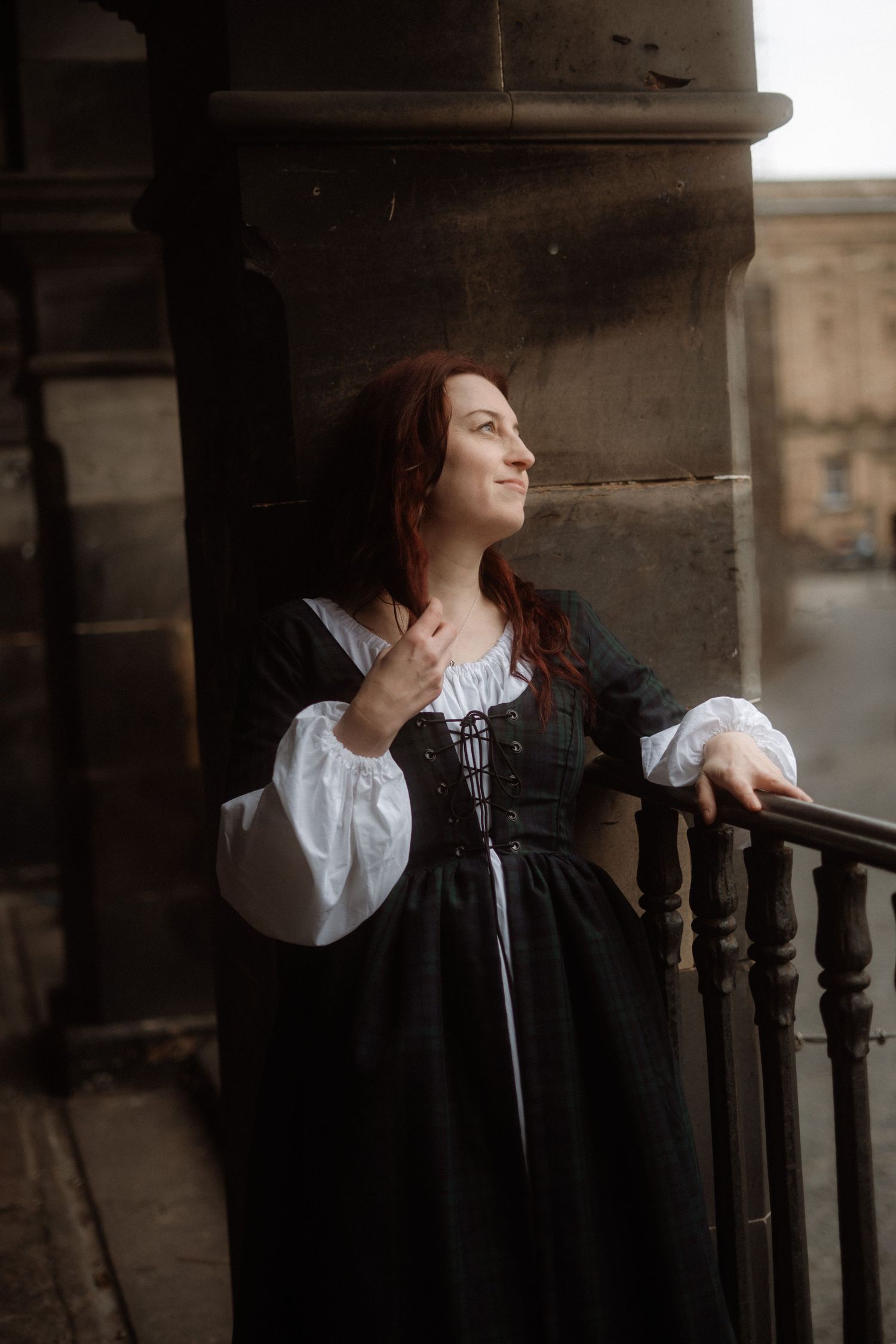 A figure in a vintage dress with white sleeves leans against dark wooden railings in a moody architectural setting.