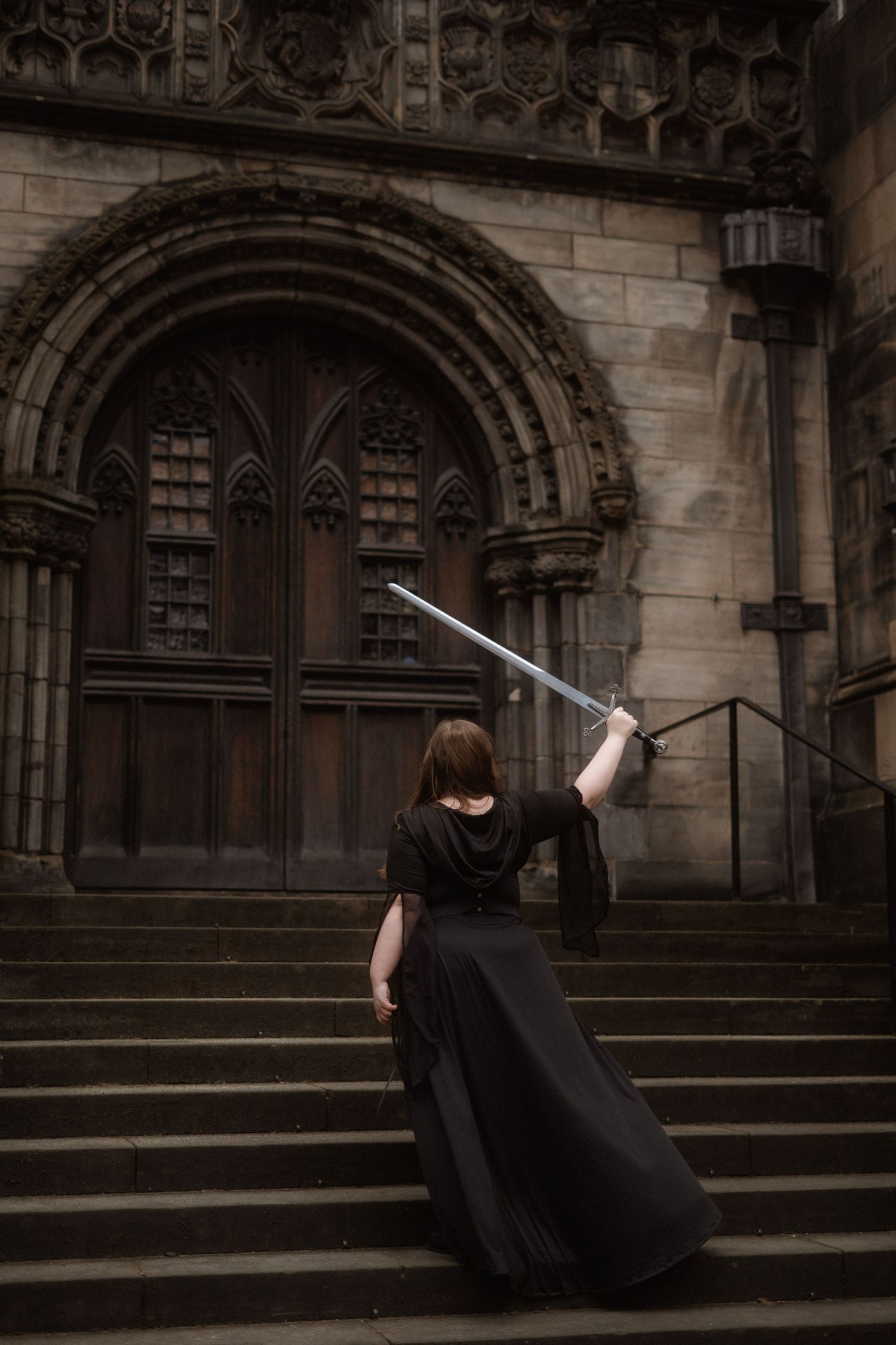 Figure in black dress holds sword while ascending gothic church steps with ornate arched doorway in background.
