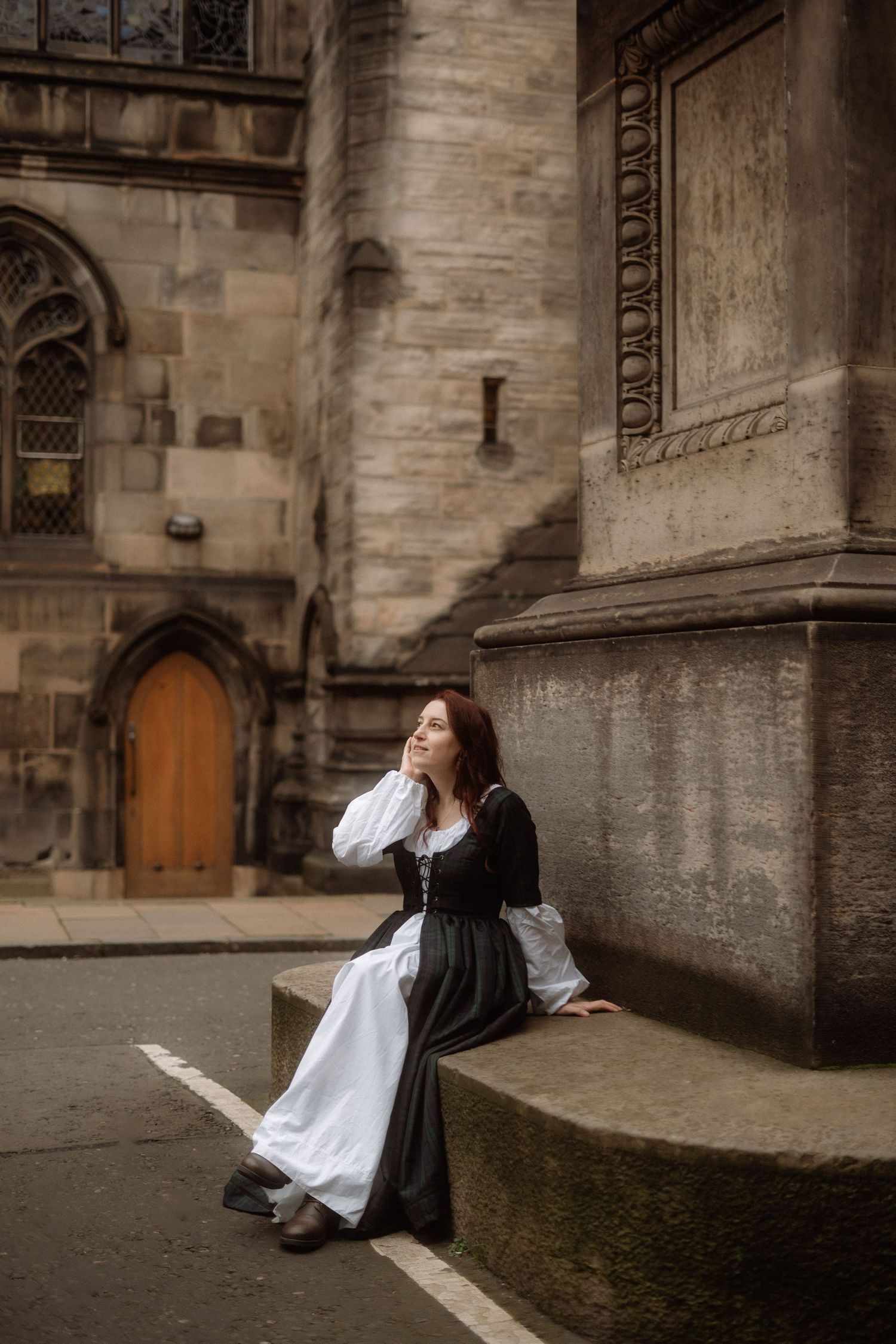 A person in black and white attire sits contemplatively against a historic stone church wall in moody lighting.