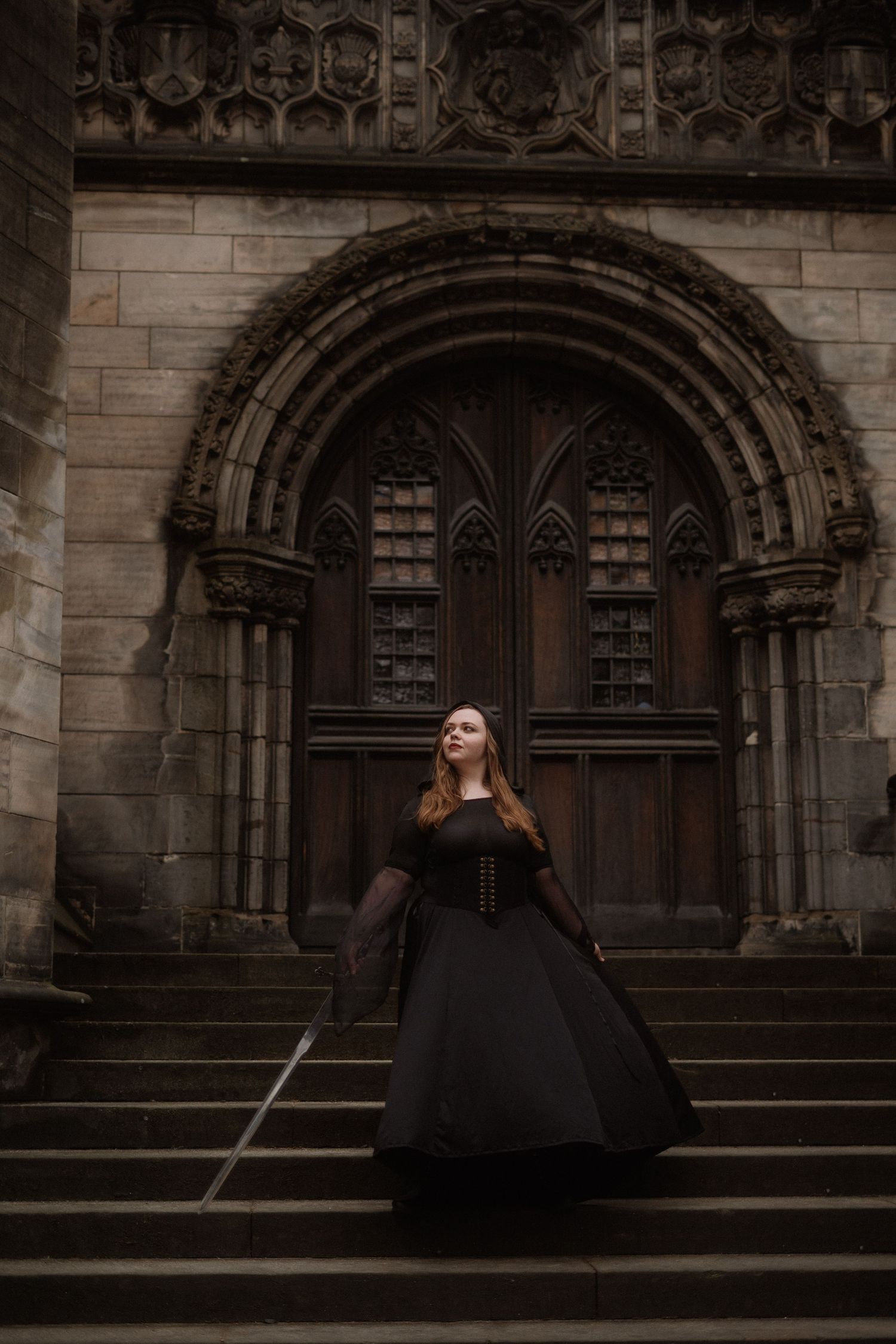 A figure in a black dress poses dramatically on stone church steps against Gothic architectural details.