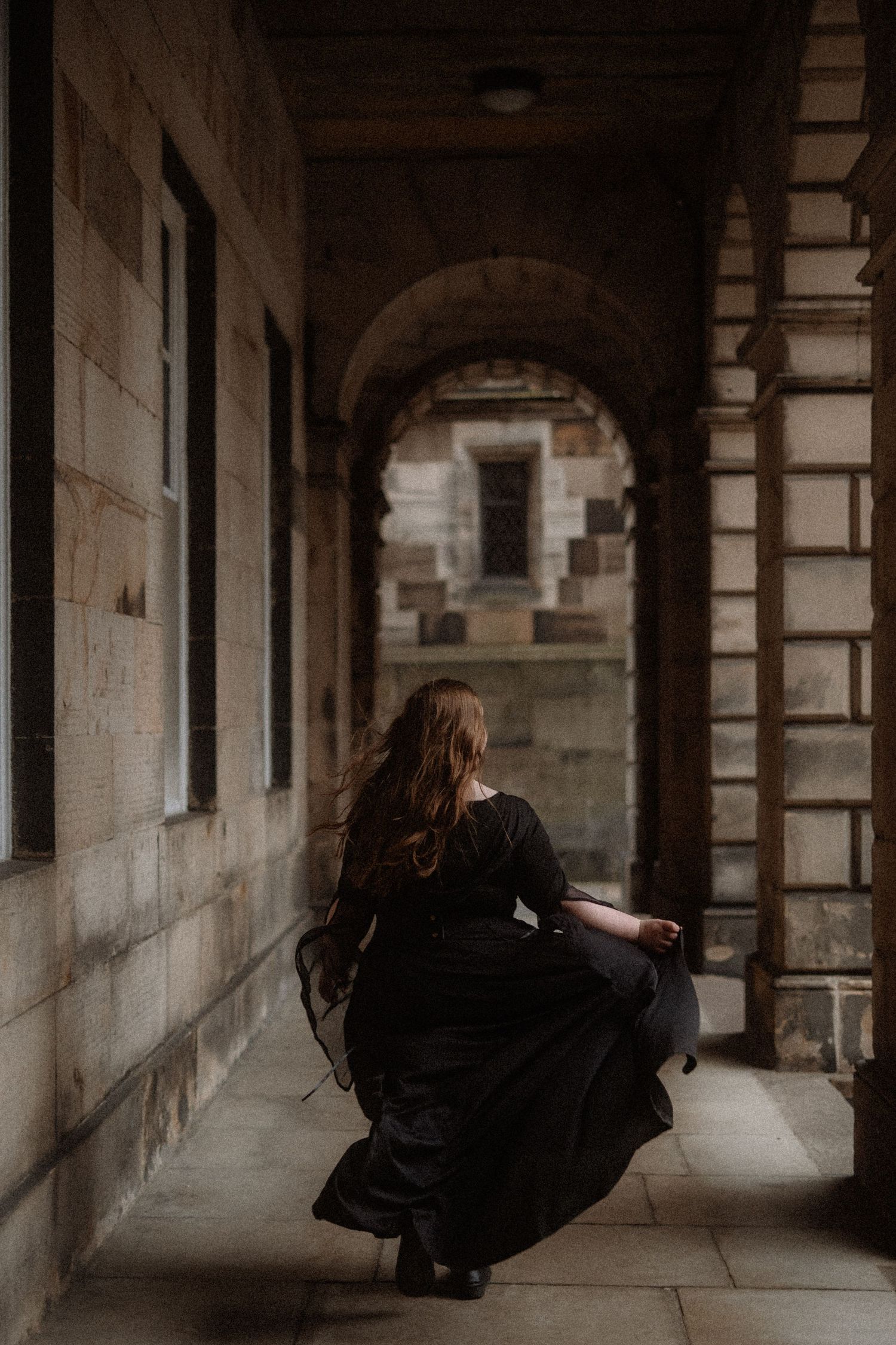 A person in dark clothing sits in a dramatic architectural corridor with arched stone columns and moody lighting.