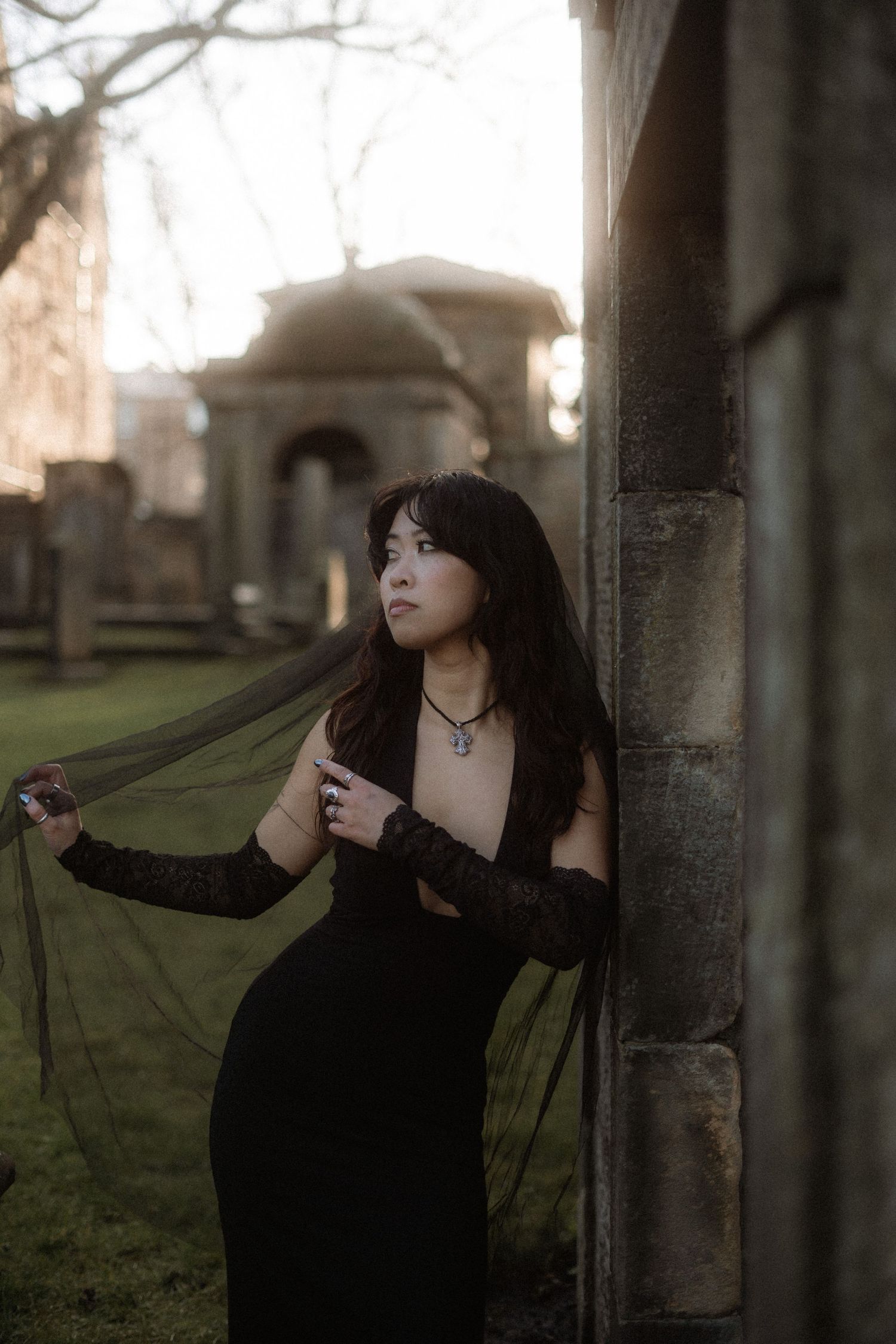 A figure in a flowing black dress stands in a moody cemetery setting at dusk, creating a gothic atmospheric scene.