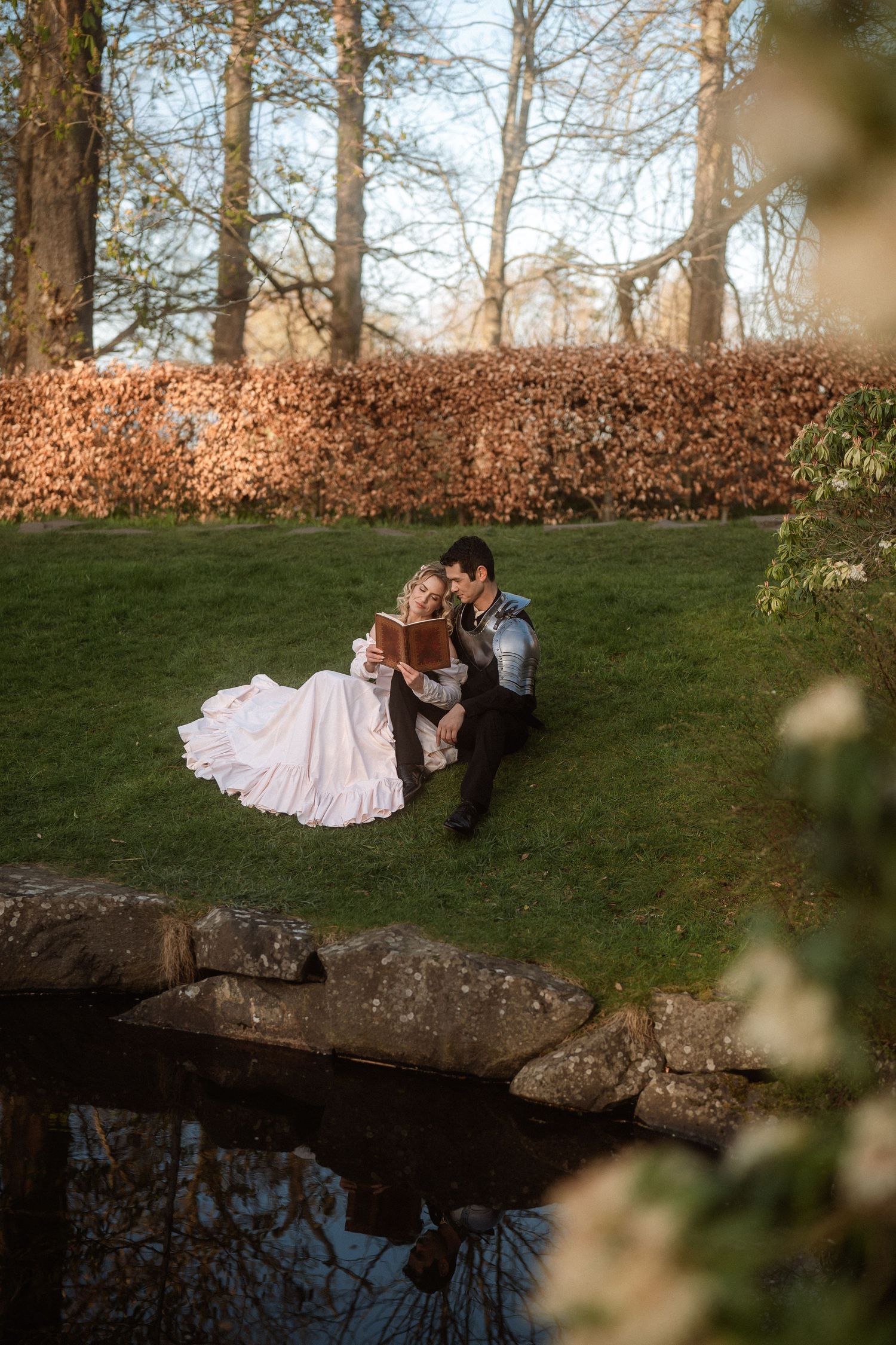 Bride and groom sitting together on green grass by a pond, surrounded by autumn trees in a romantic outdoor wedding setting.