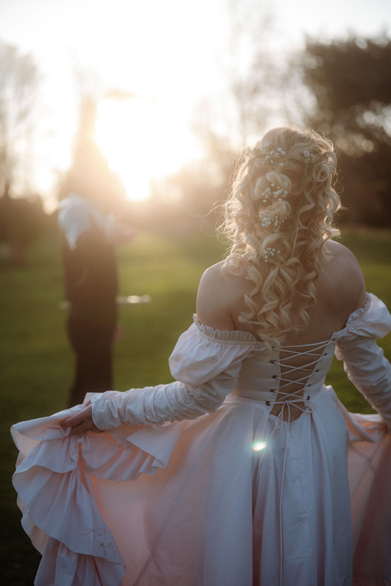 Blonde woman in elegant lace-up corset gown with curly hair, photographed at golden sunset in outdoor garden setting.