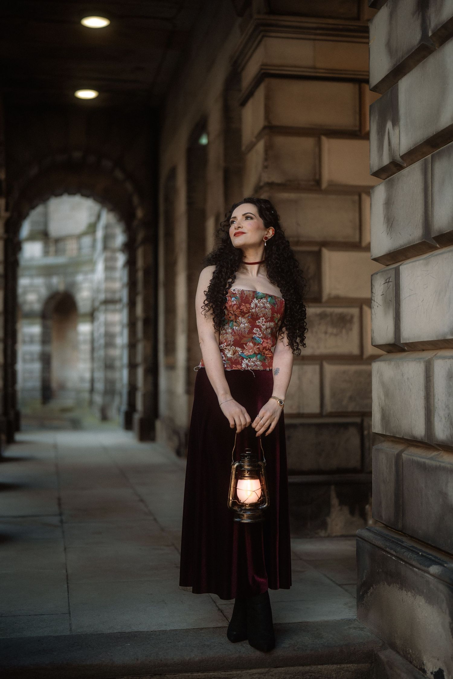 Woman in floral corset and burgundy velvet skirt holds a glowing lantern beside stone archway at dusk.