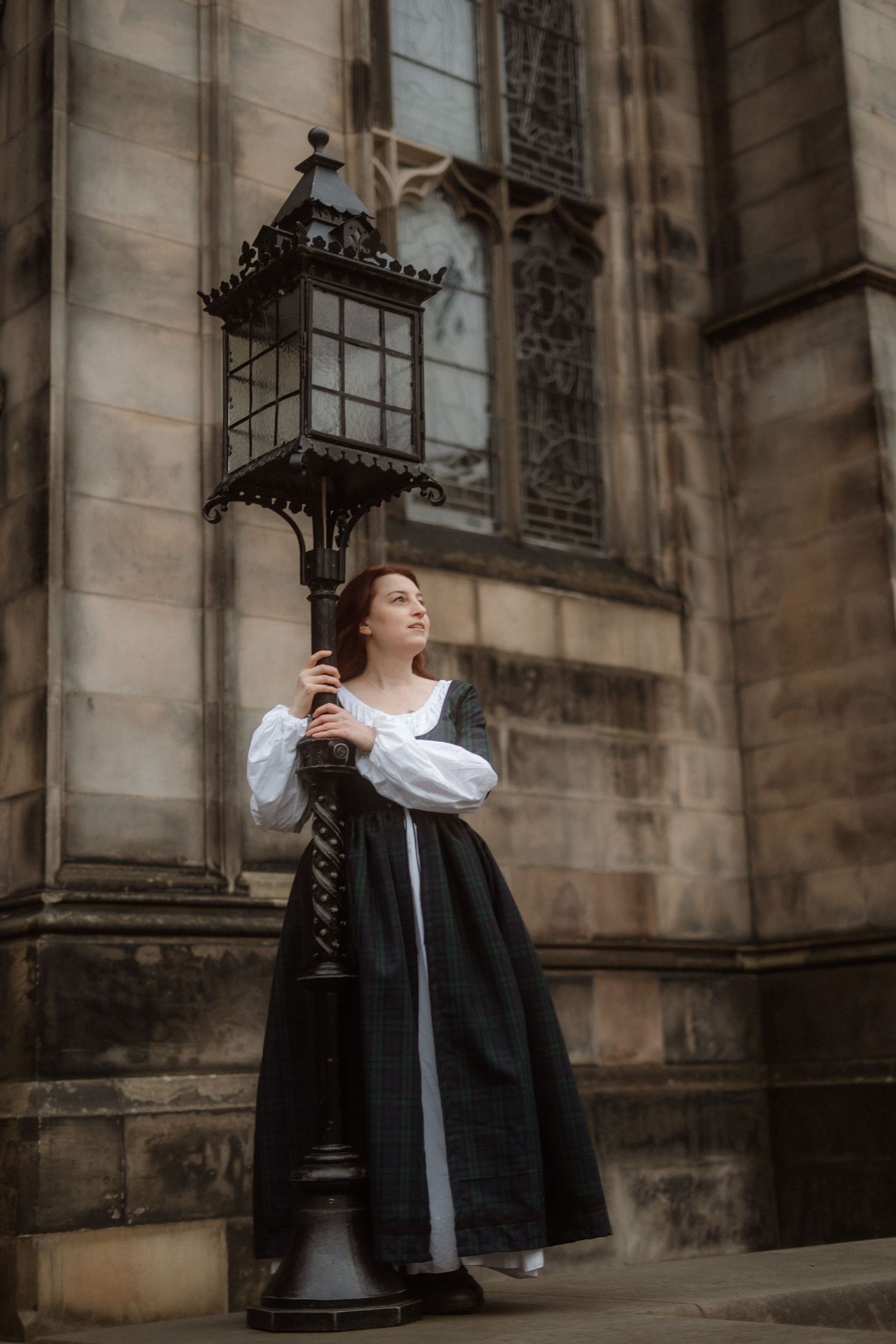 A person in a black dress with white collar stands against a stone church wall with vintage lanterns.