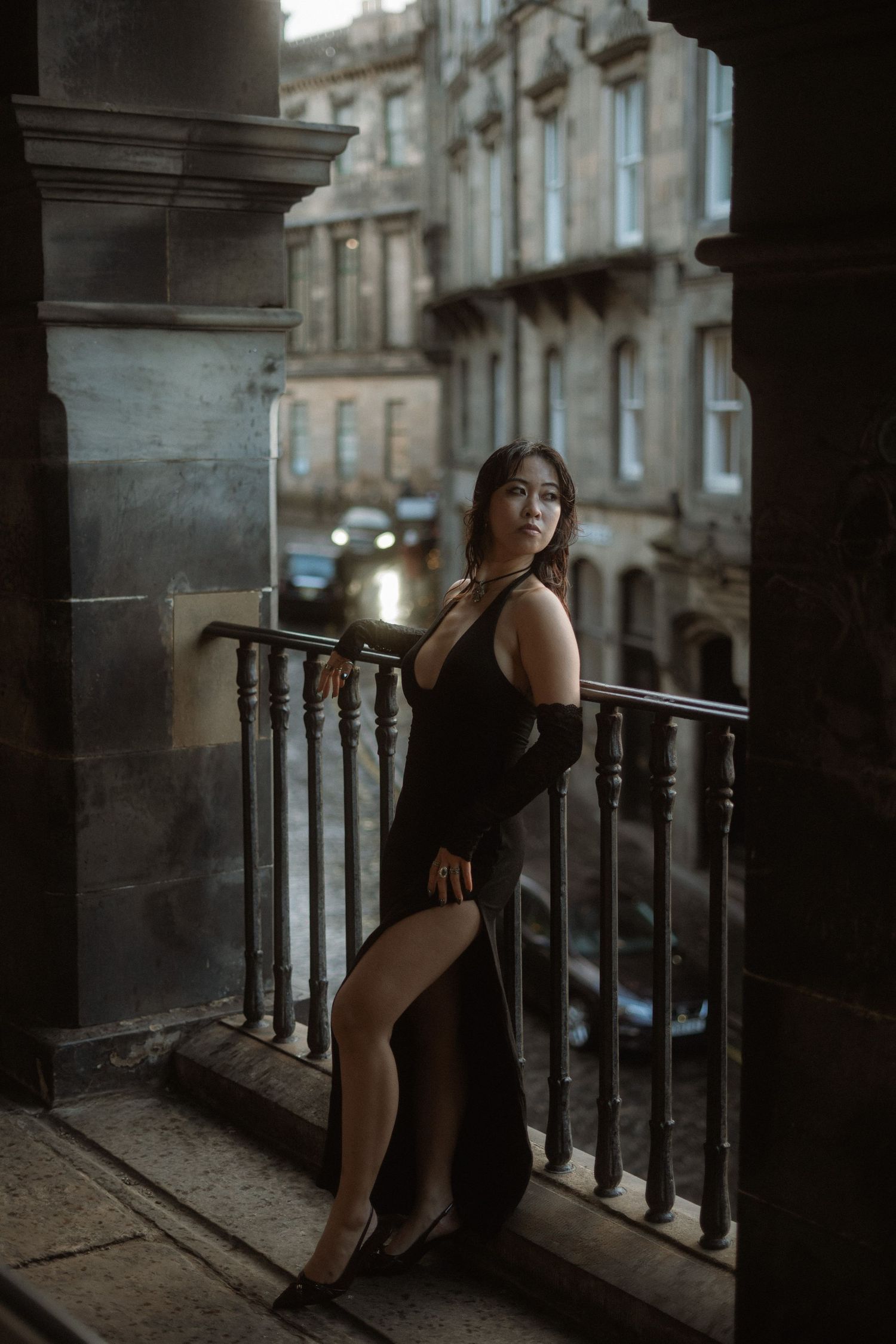 A woman in a black dress poses dramatically on a vintage balcony in moody lighting.