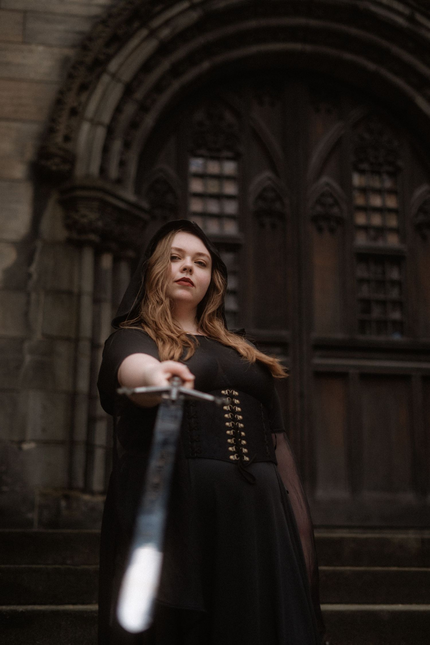 A person in a black dress stands against a gothic architectural backdrop while holding a glowing light source.
