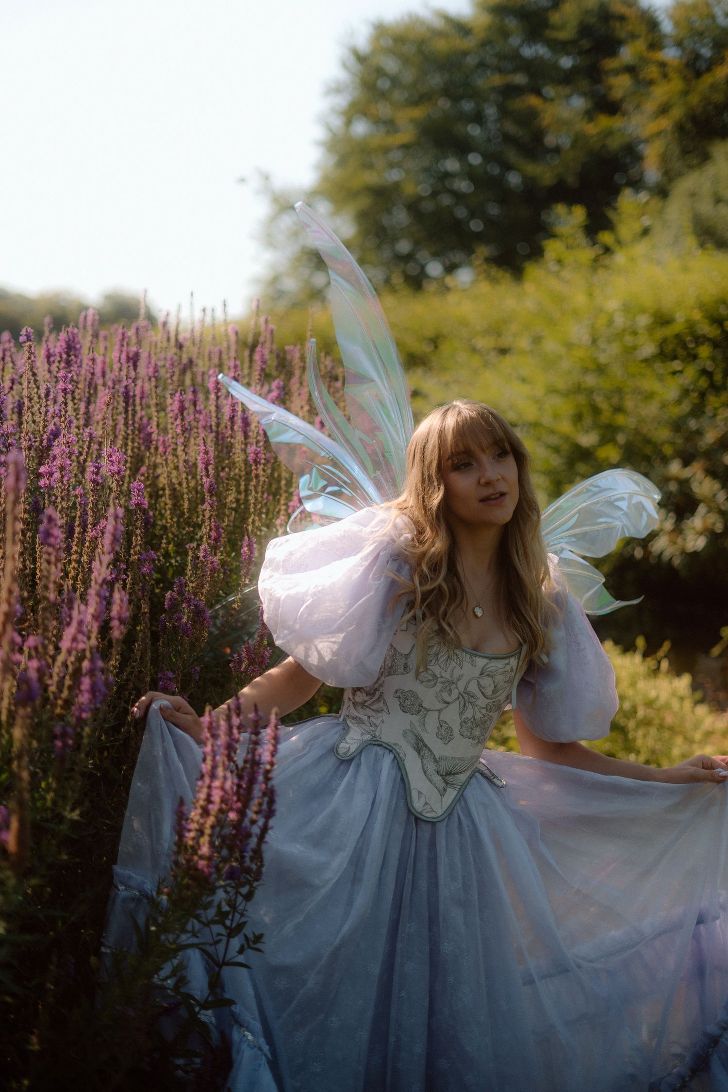 A fairy-like figure in a blue dress and wings twirls gracefully in a garden with purple flowers in the background.