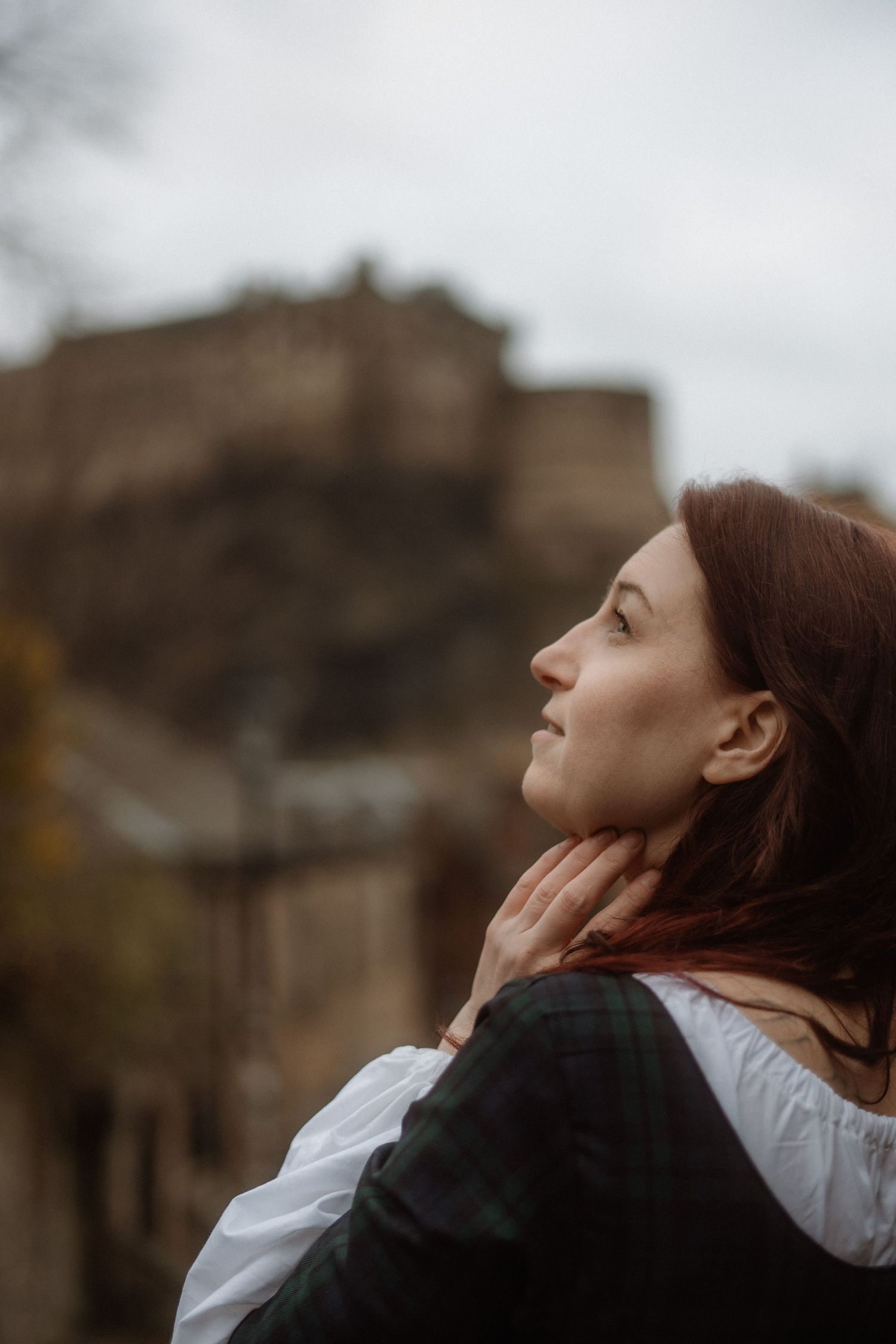 A person in period dress gazes upward with Edinburgh Castle visible in the background on an overcast day.