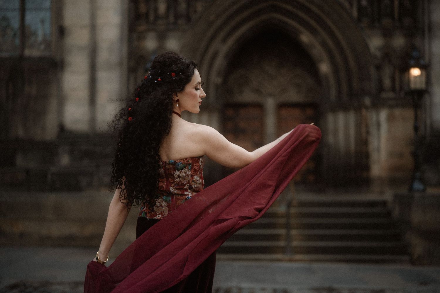 Woman in burgundy gown with flowing fabric poses before gothic cathedral architecture.