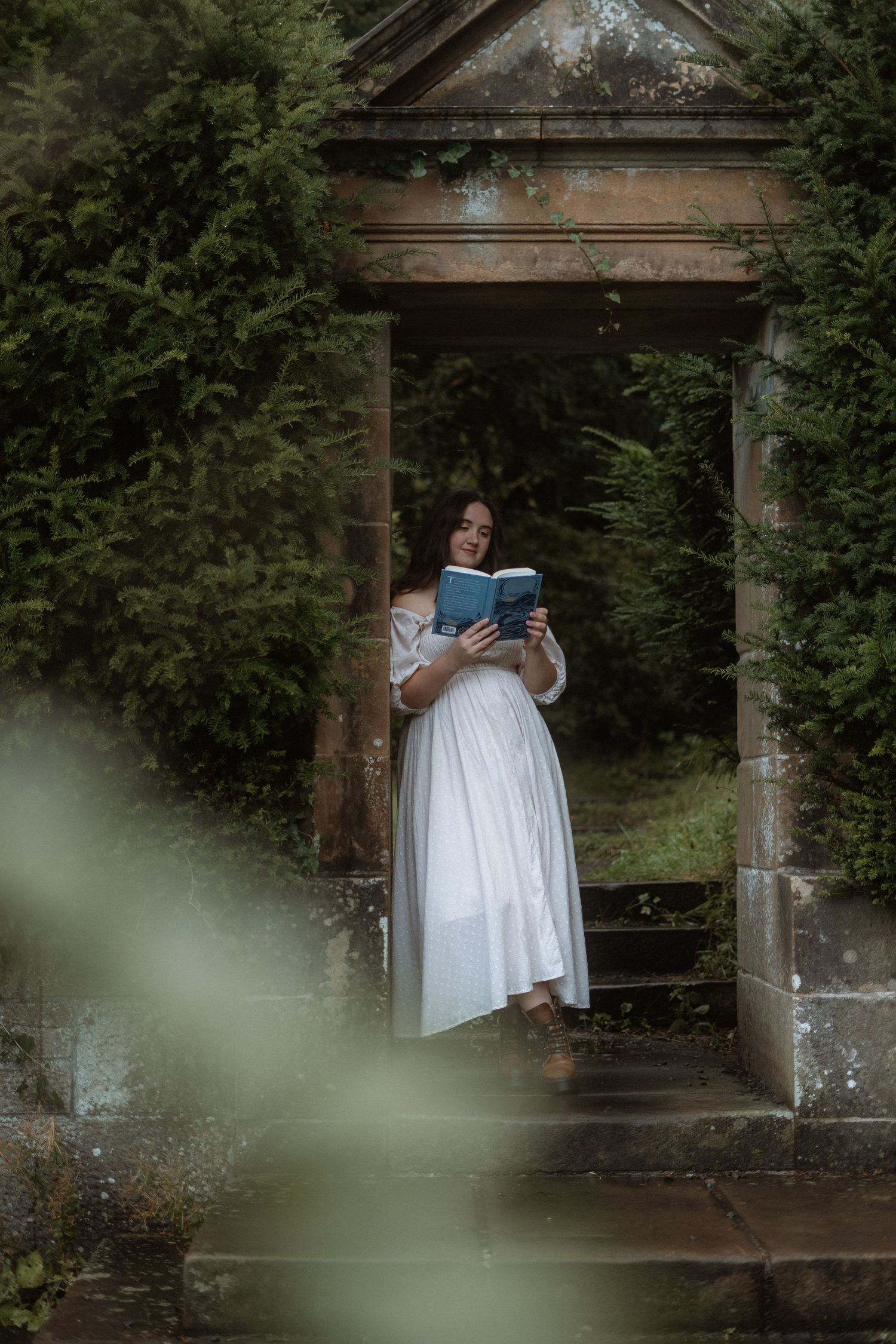 A figure in white reads a book under an ornate stone archway in a garden.