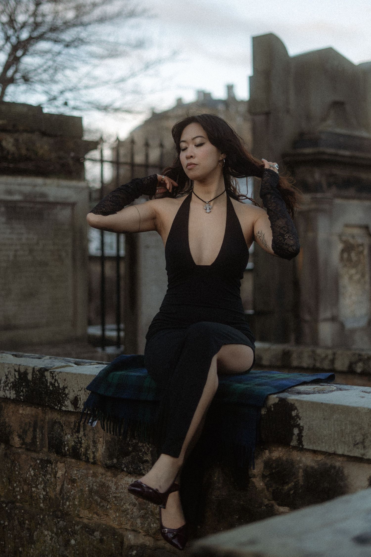 A woman in a black halter dress sits on a stone wall in a moody cemetery setting at dusk.