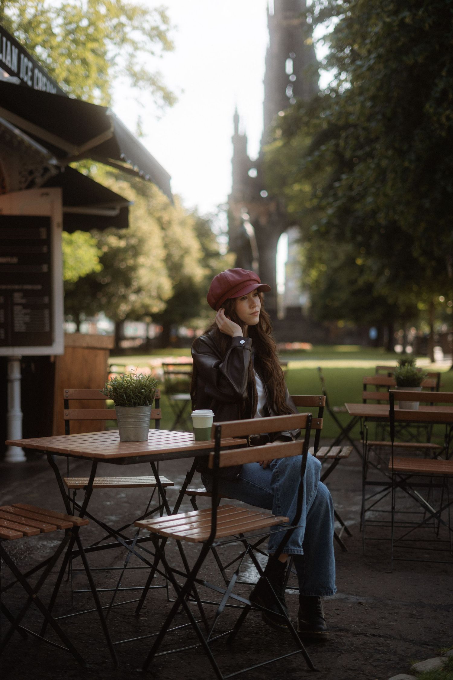 Woman in red cap and leather jacket sits at outdoor cafe table with coffee, green park and Gothic tower behind her.
