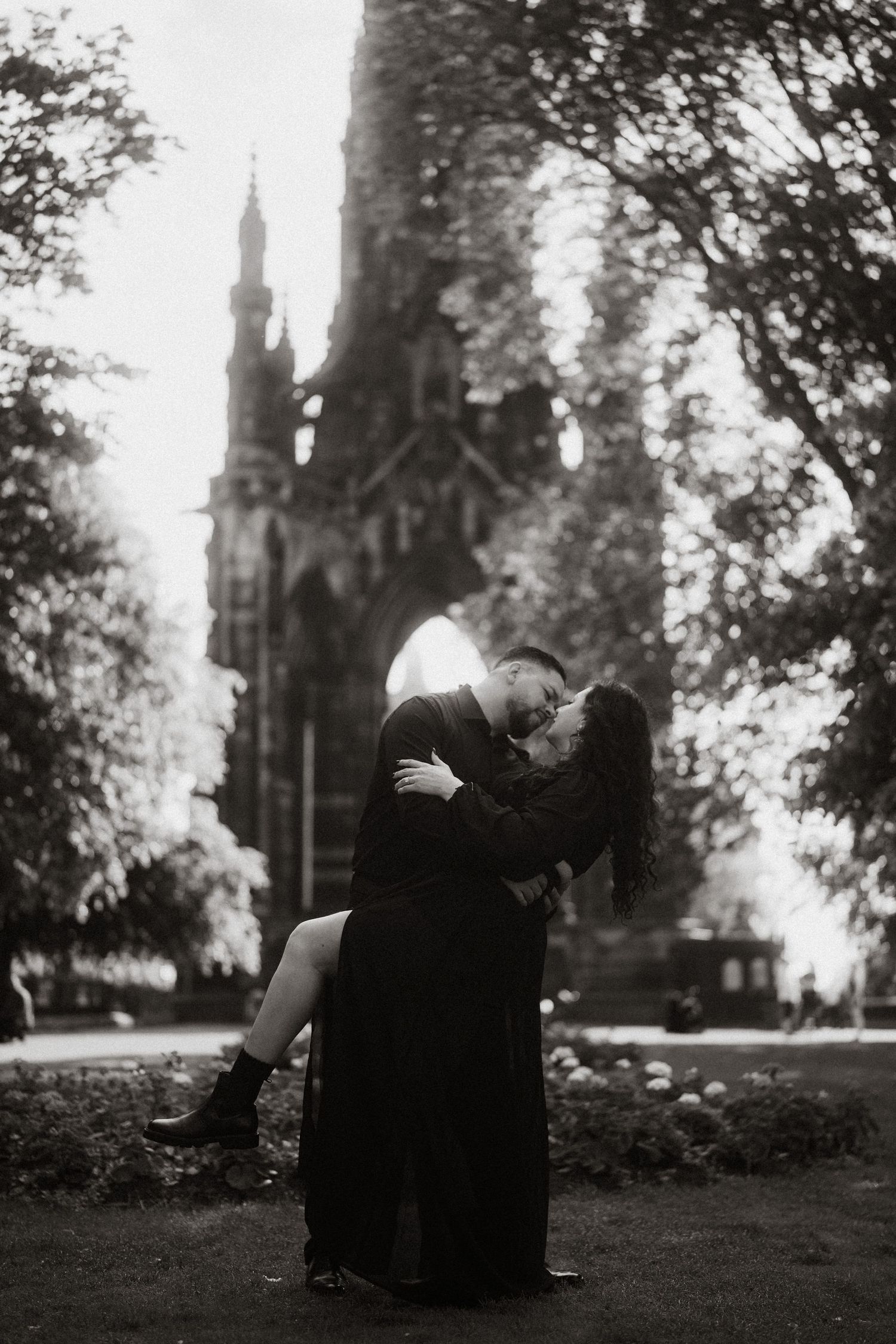 Black and white photo of a couple kissing in front of a Gothic monument surrounded by trees.