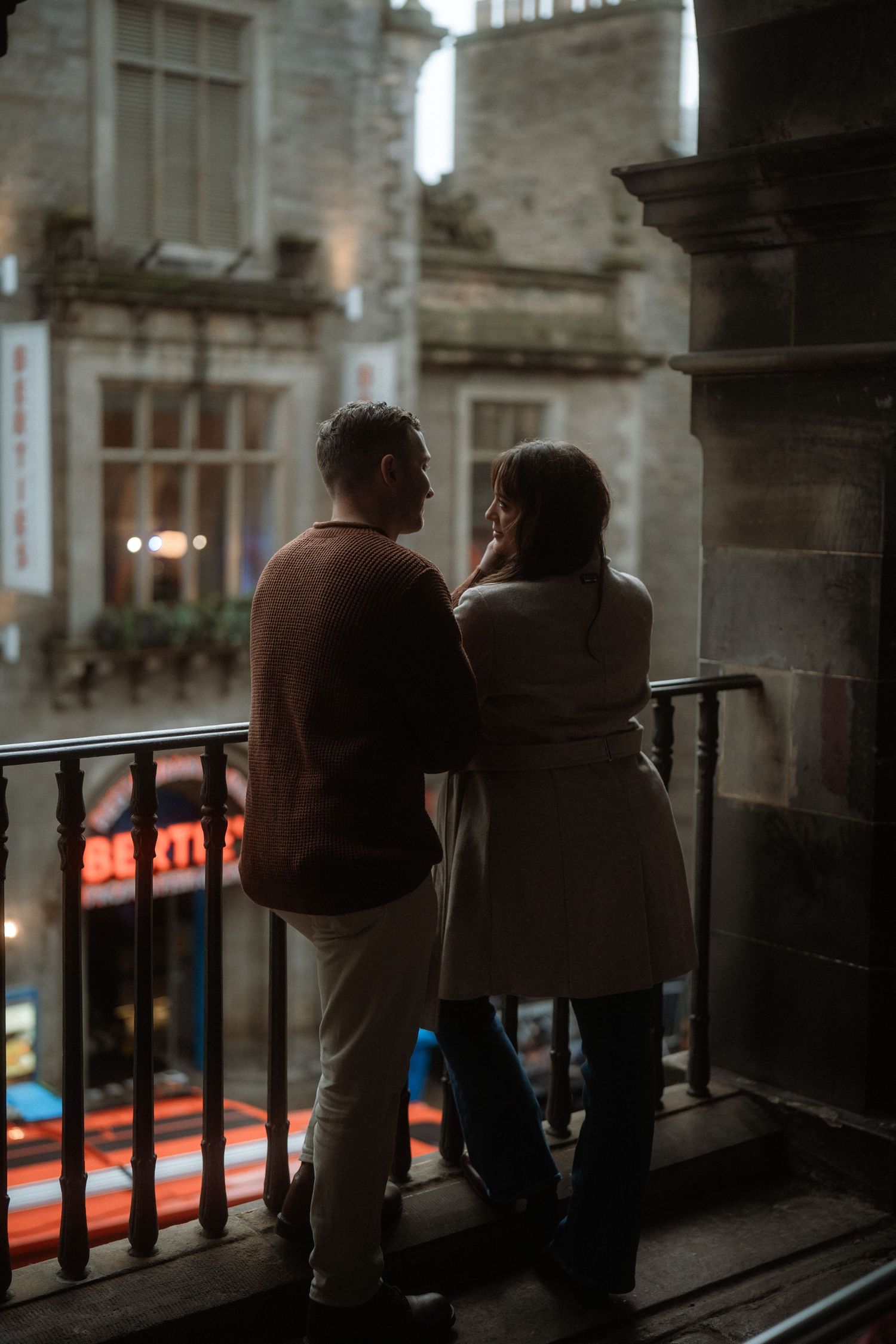 Romantic couple standing on a dimly lit balcony overlooking historic stone buildings at dusk.