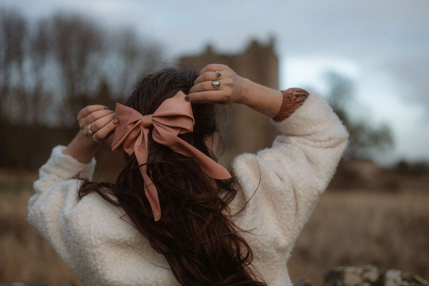A figure in a cozy cream sweater stands in a field at dusk with a large pink bow in their dark flowing hair.