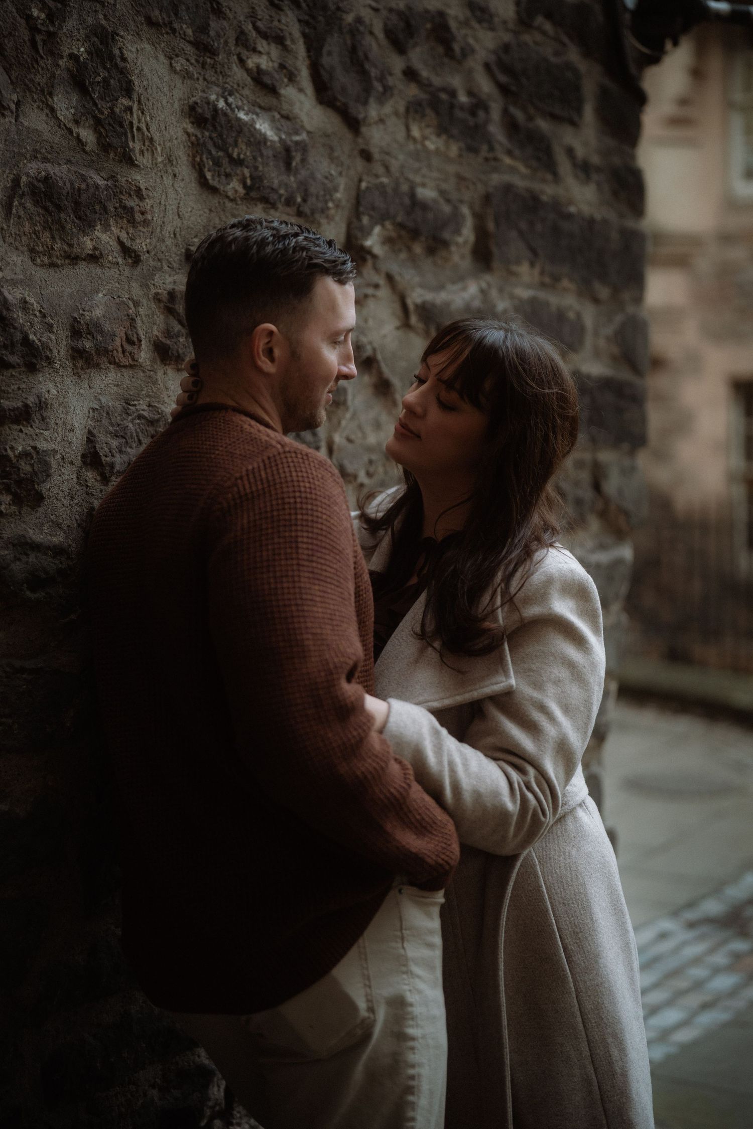 Romantic couple embracing against a stone wall, woman in cream coat gazing up at man in brown sweater.