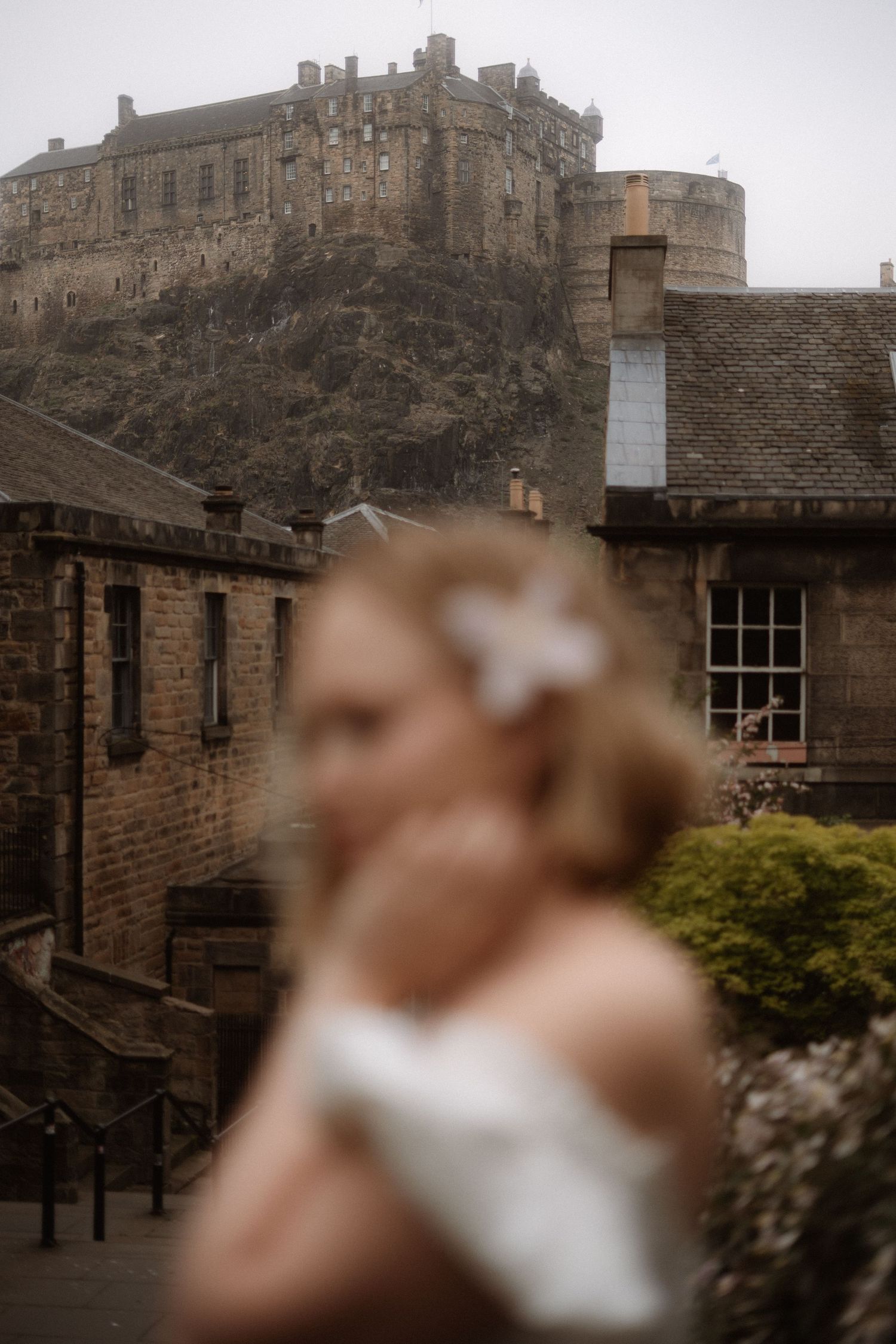Blurred bride with flower hair accessory in foreground, Edinburgh Castle on misty hilltop behind stone buildings.