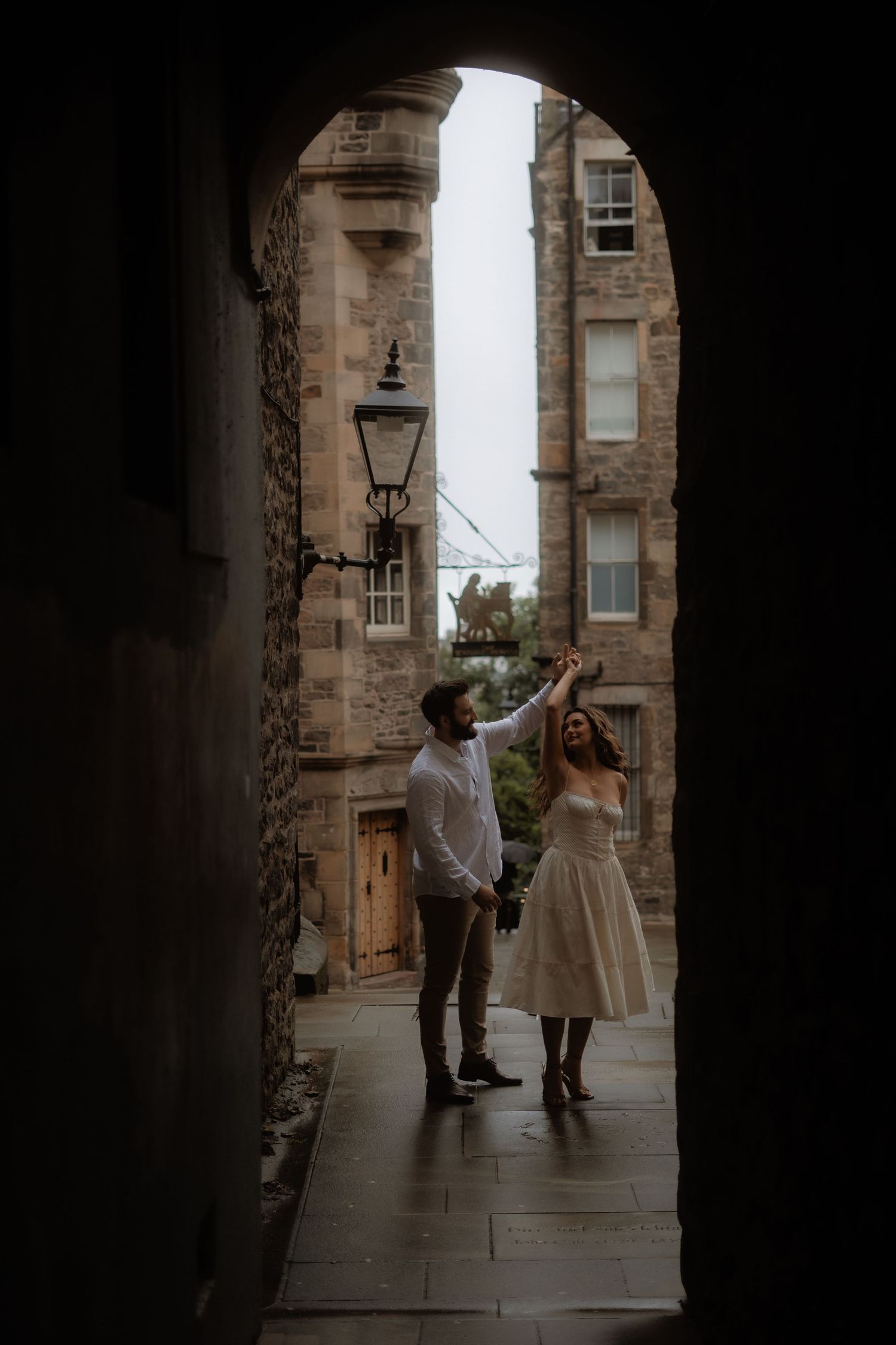 Couple dancing under a stone archway in Edinburgh's Old Town, with historic buildings and a vintage lamp post in the background.