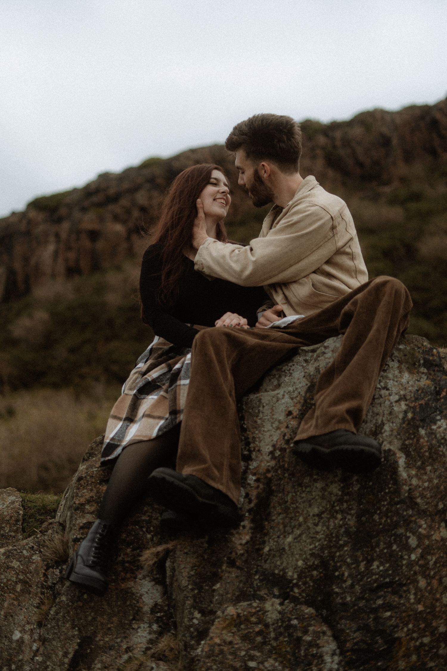 A couple shares a tender moment while sitting on rocky terrain against a moody landscape.