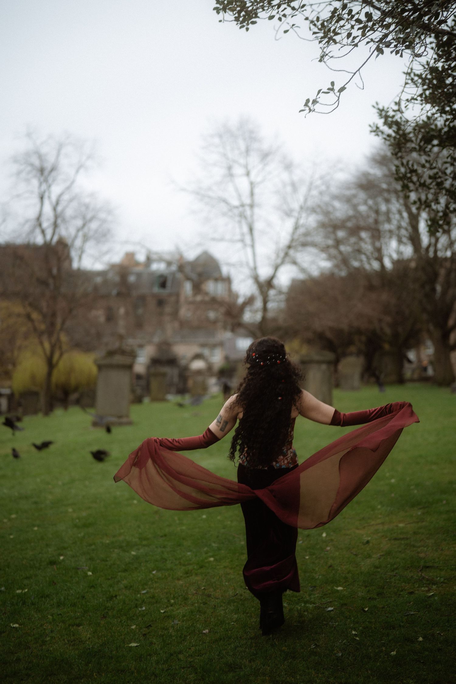 Woman in flowing red gown dancing in a historic cemetery with ravens and Gothic architecture in the background.