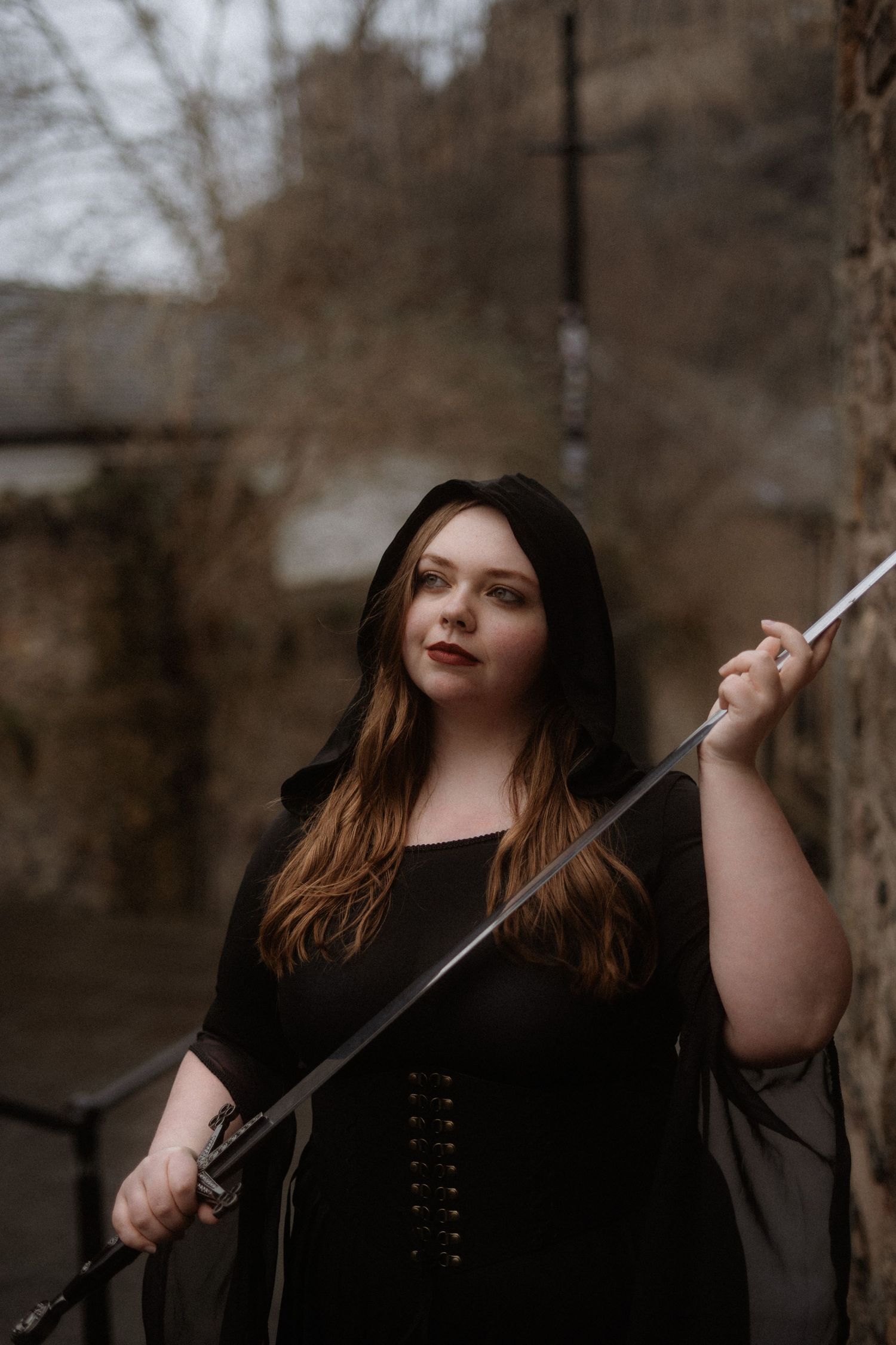 A person in dark clothing poses dramatically in a moody forest setting with artistic lighting and shadows.