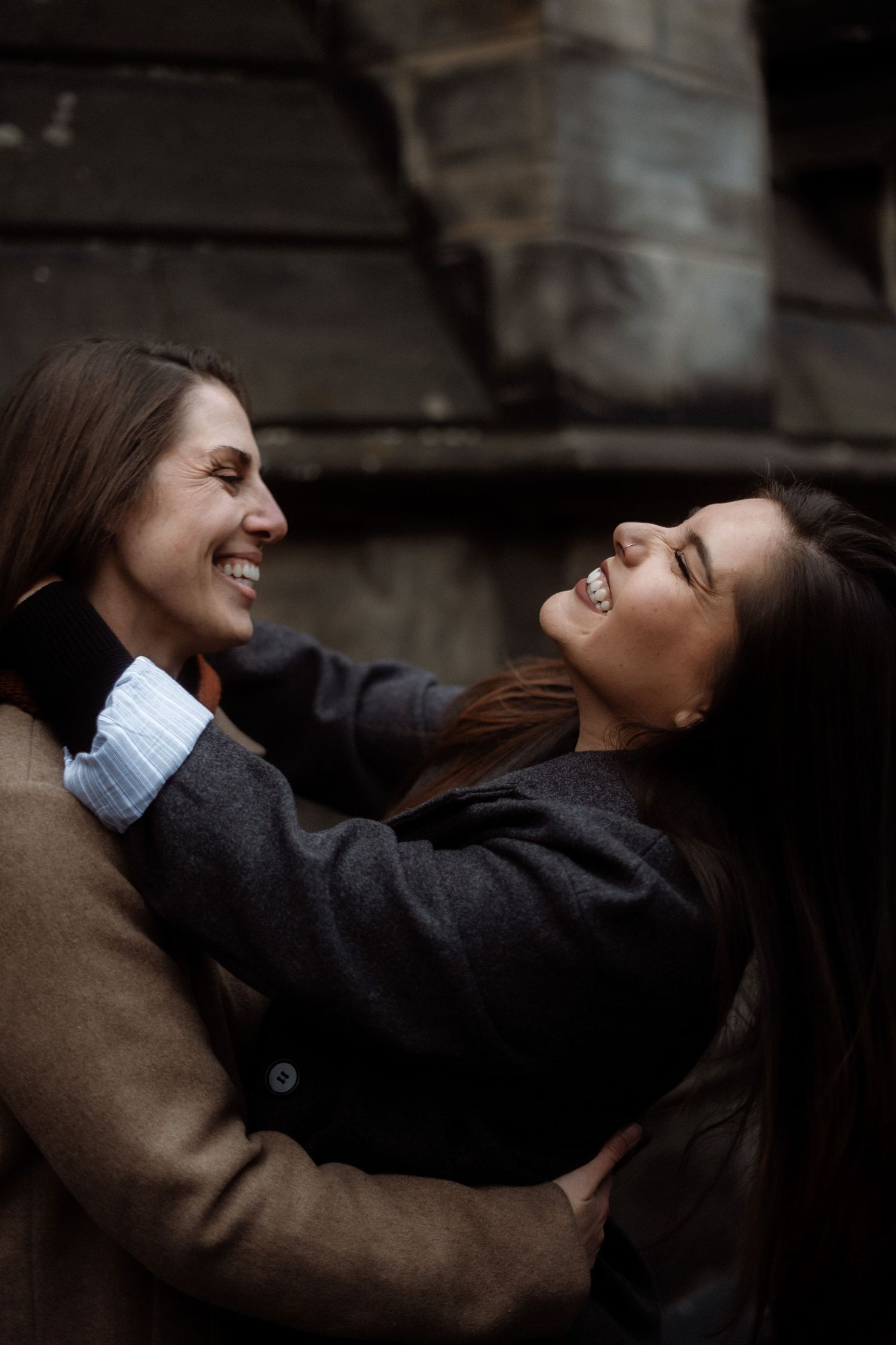 Two people share a joyful embrace and laugh together while wearing winter coats outdoors in Edinburgh, Scotland.