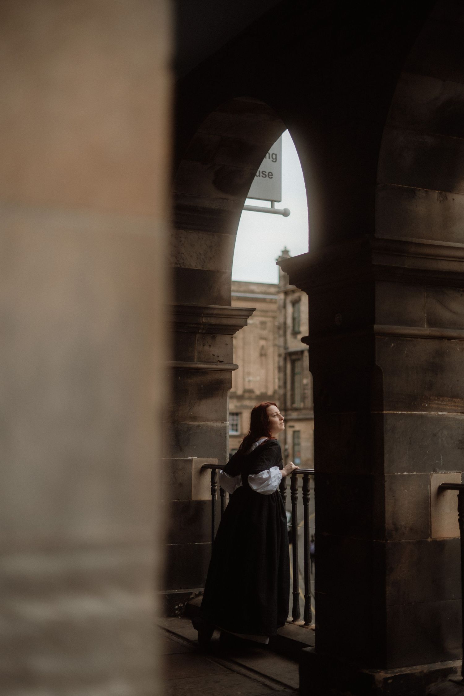 Person in black clothing stands in shadows beneath historic stone archways with dramatic lighting.