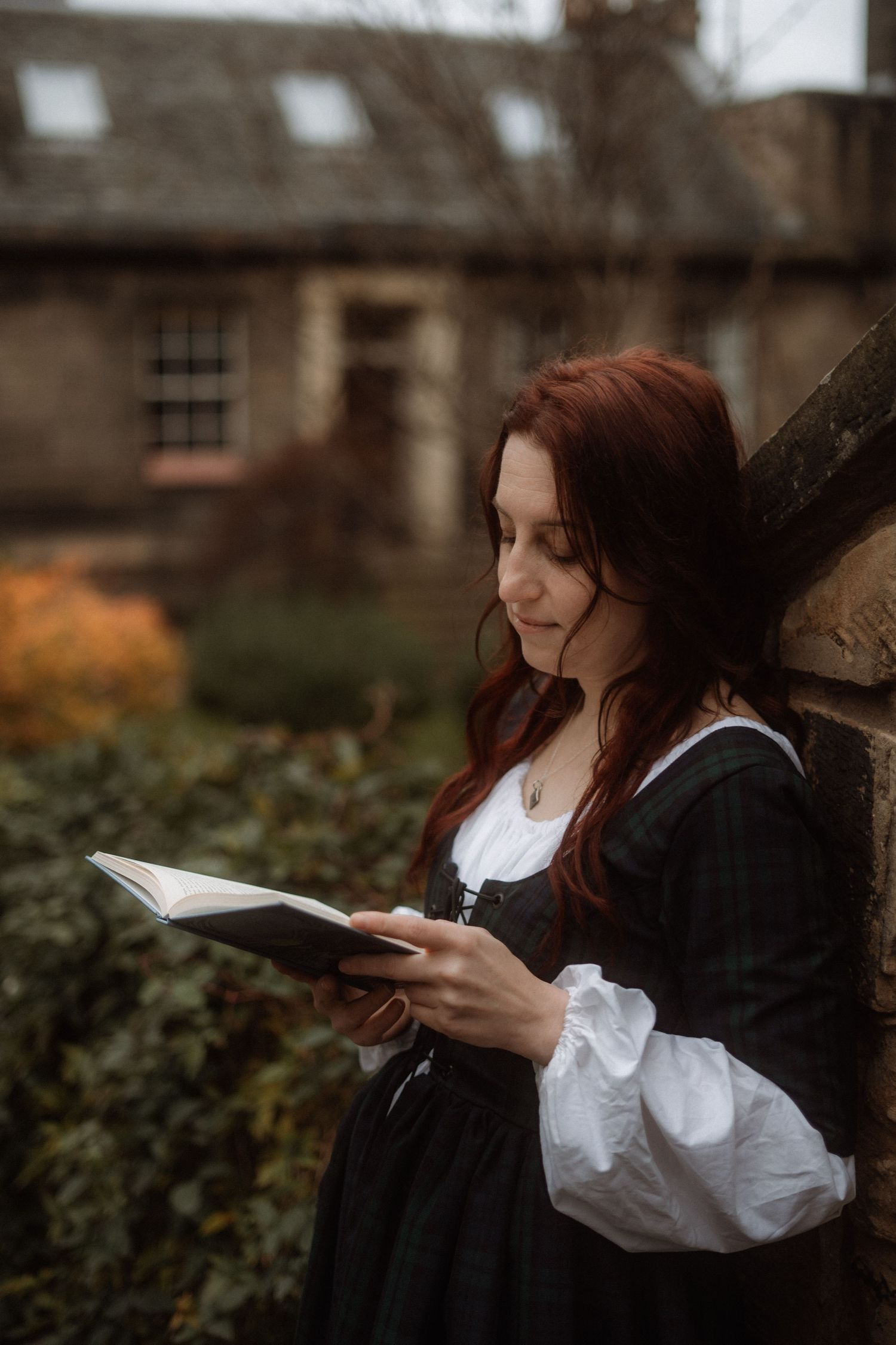 A woman in a vintage dress reads a book in a garden with autumn foliage and historic buildings in the background.