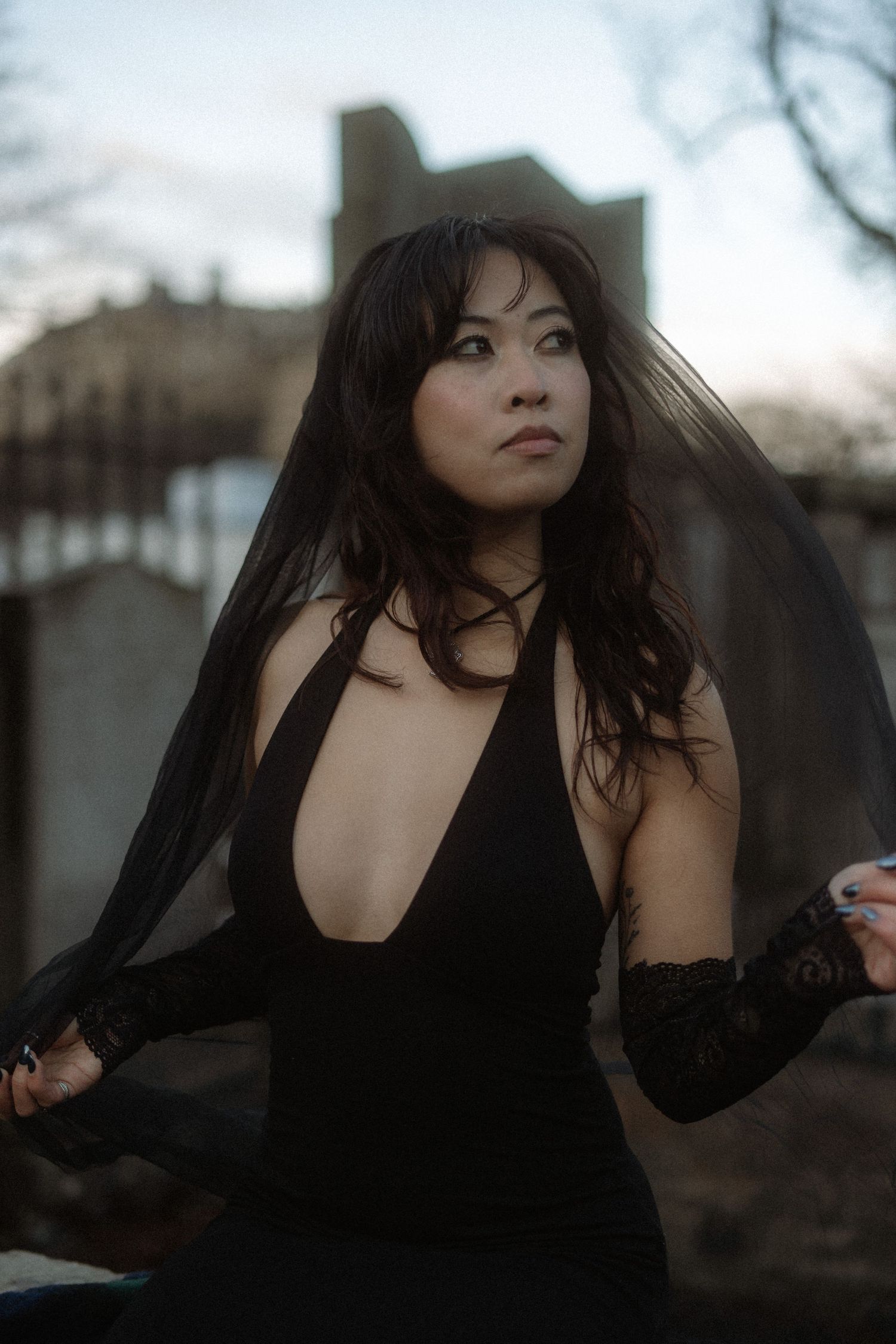Dramatic portrait of someone in a flowing black dress with plunging neckline against a moody outdoor background.