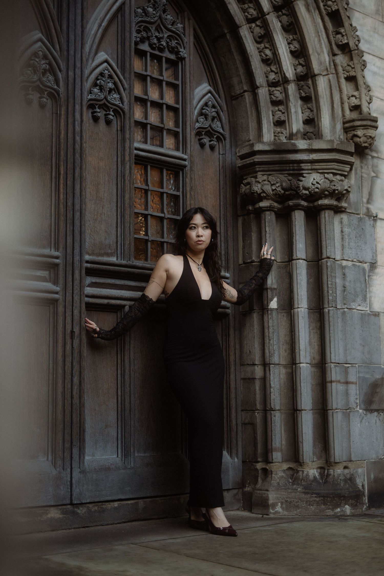 A person in a black dress poses dramatically against ornate gothic stone archways and columns.