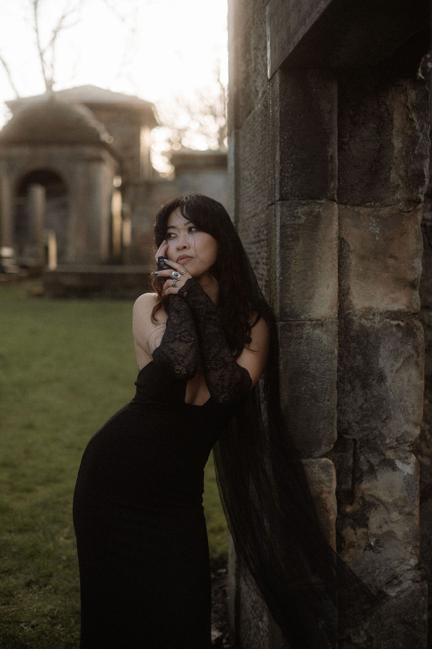 A person in a black dress poses dramatically between stone pillars at dusk in an artistic outdoor photoshoot.