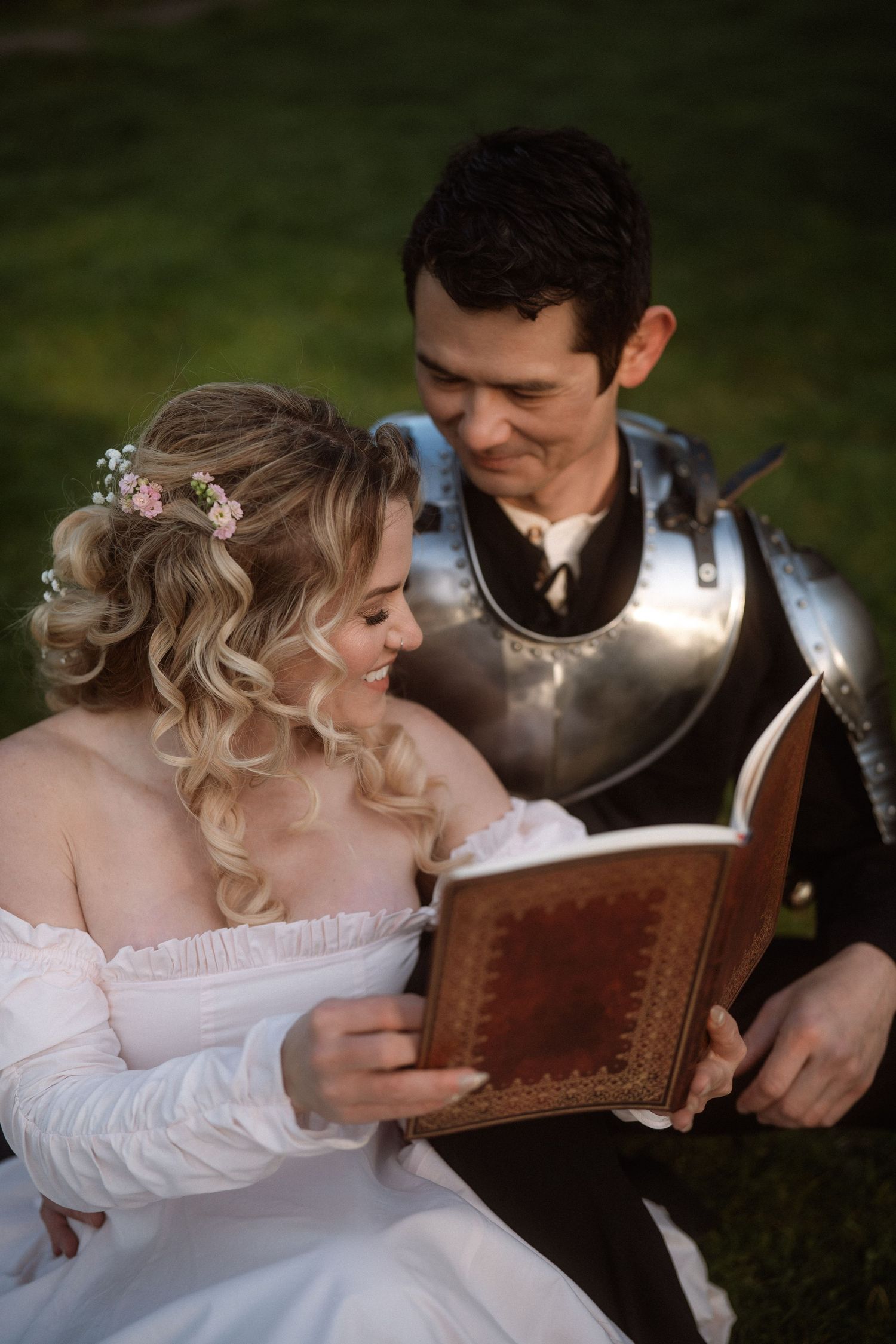 A bride in white dress and groom in medieval armor sit together reading an antique book outdoors.