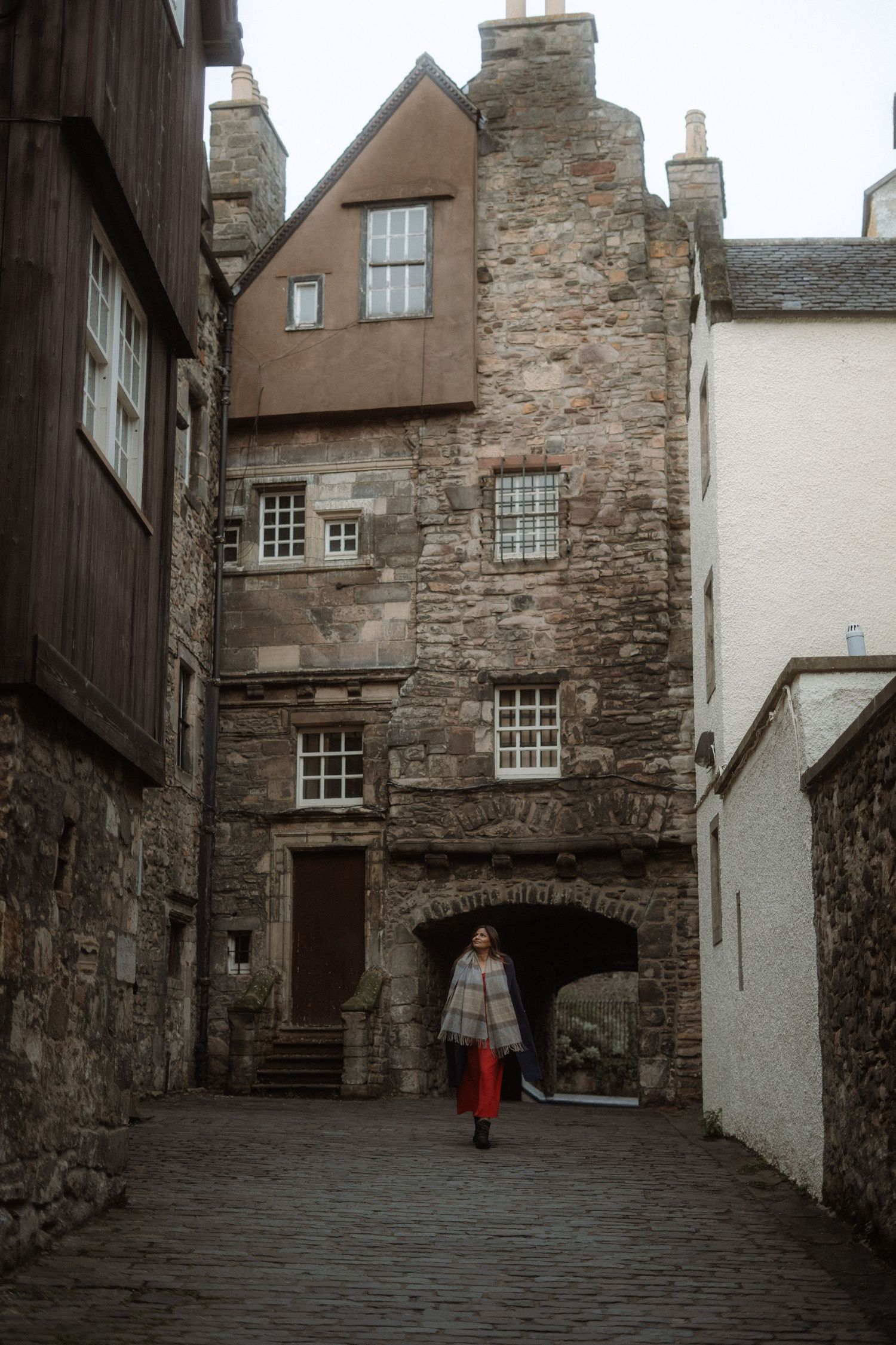 Woman walking through historic stone alleyway in Edinburgh Old Town, Scotland, wearing plaid coat and red trousers.