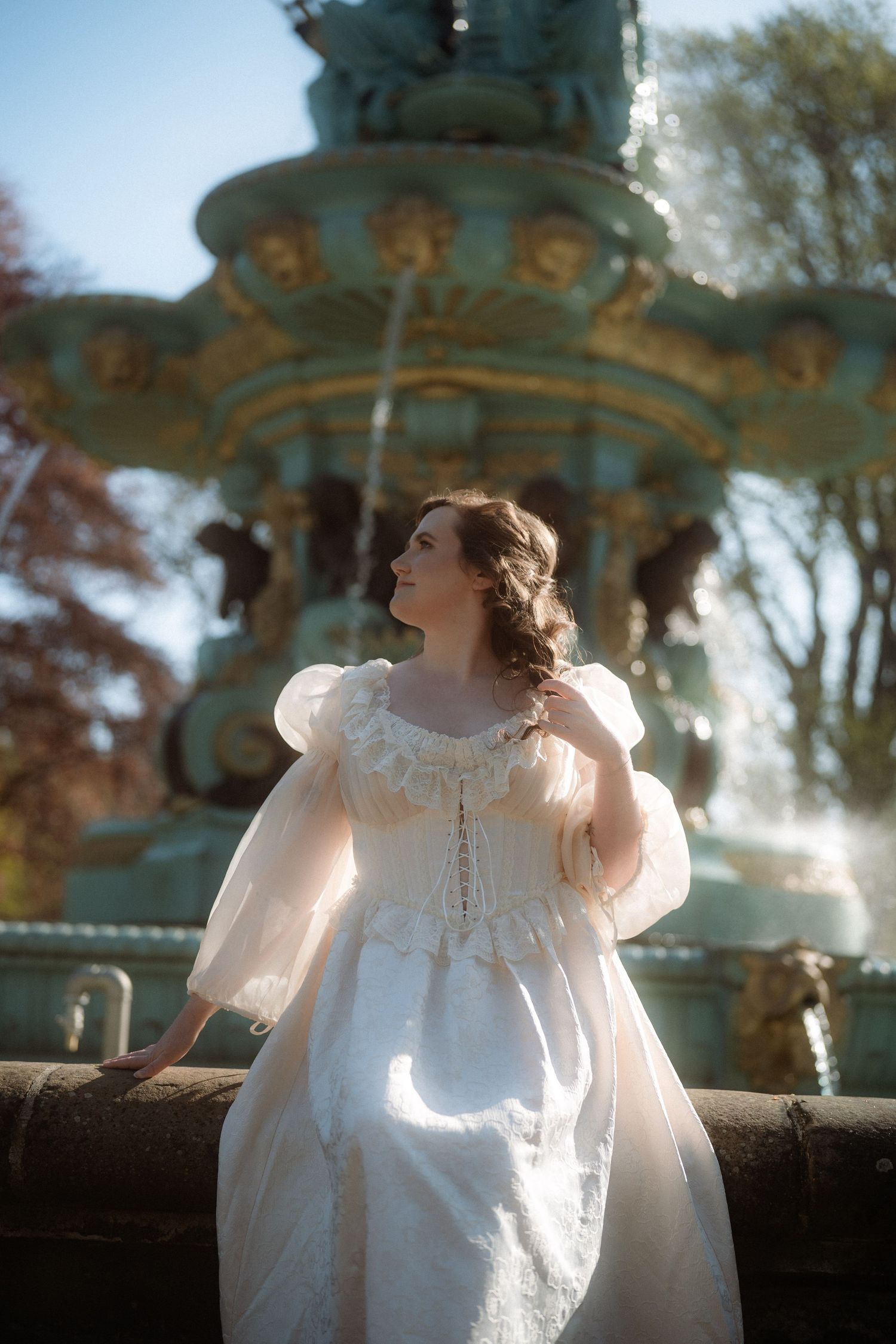 Woman in vintage white gown poses gracefully by ornate fountain in sunlit park.
