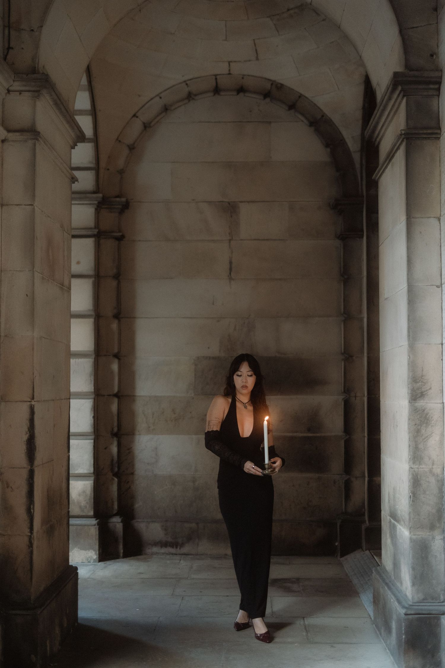 A figure in a black dress holds a candle while standing in a stone archway of a dimly lit historic building.