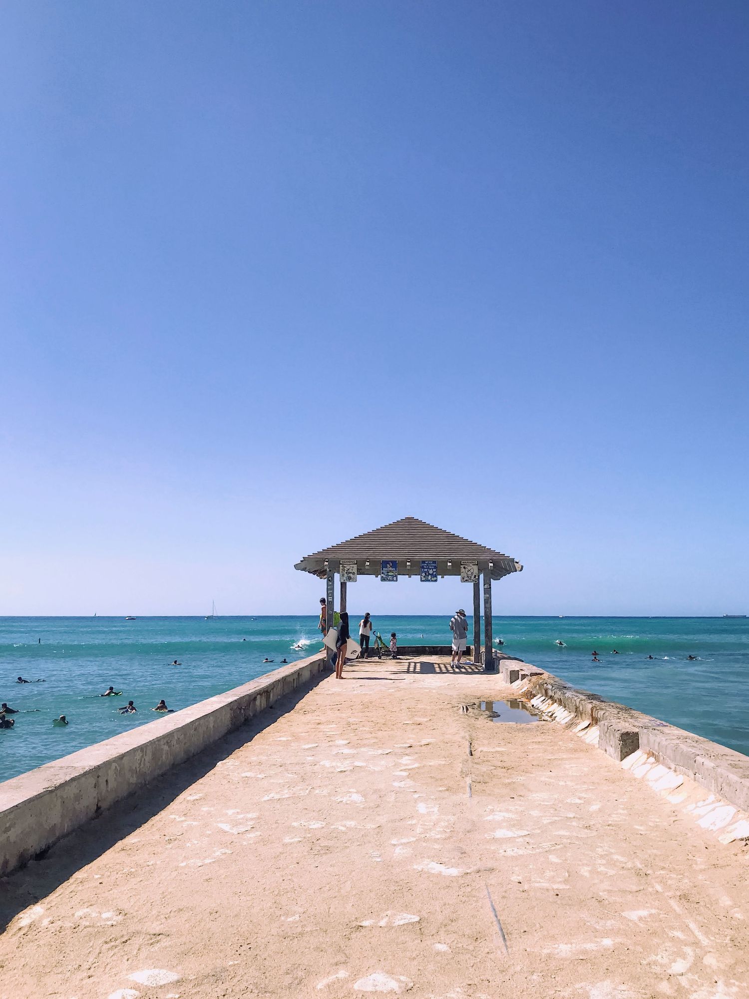 A sunny Hawaii beach scene with people walking along a concrete pier, swimmers in bright blue ocean water, and a pavilion at the end.