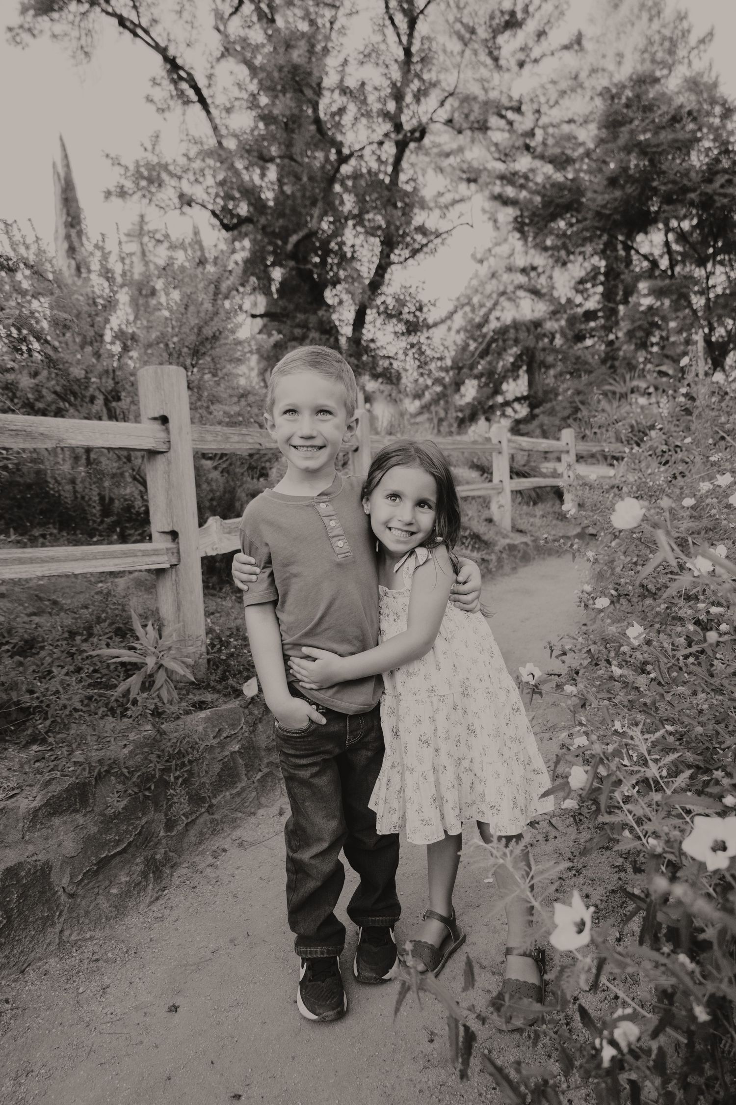 Black and white photo of two young siblings hugging on a garden path near a wooden fence.