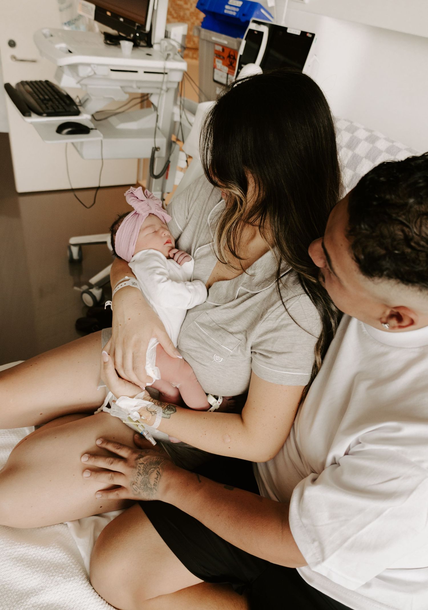 New parents in hospital room tenderly holding their newborn baby dressed in pink, sharing a precious first bonding moment.