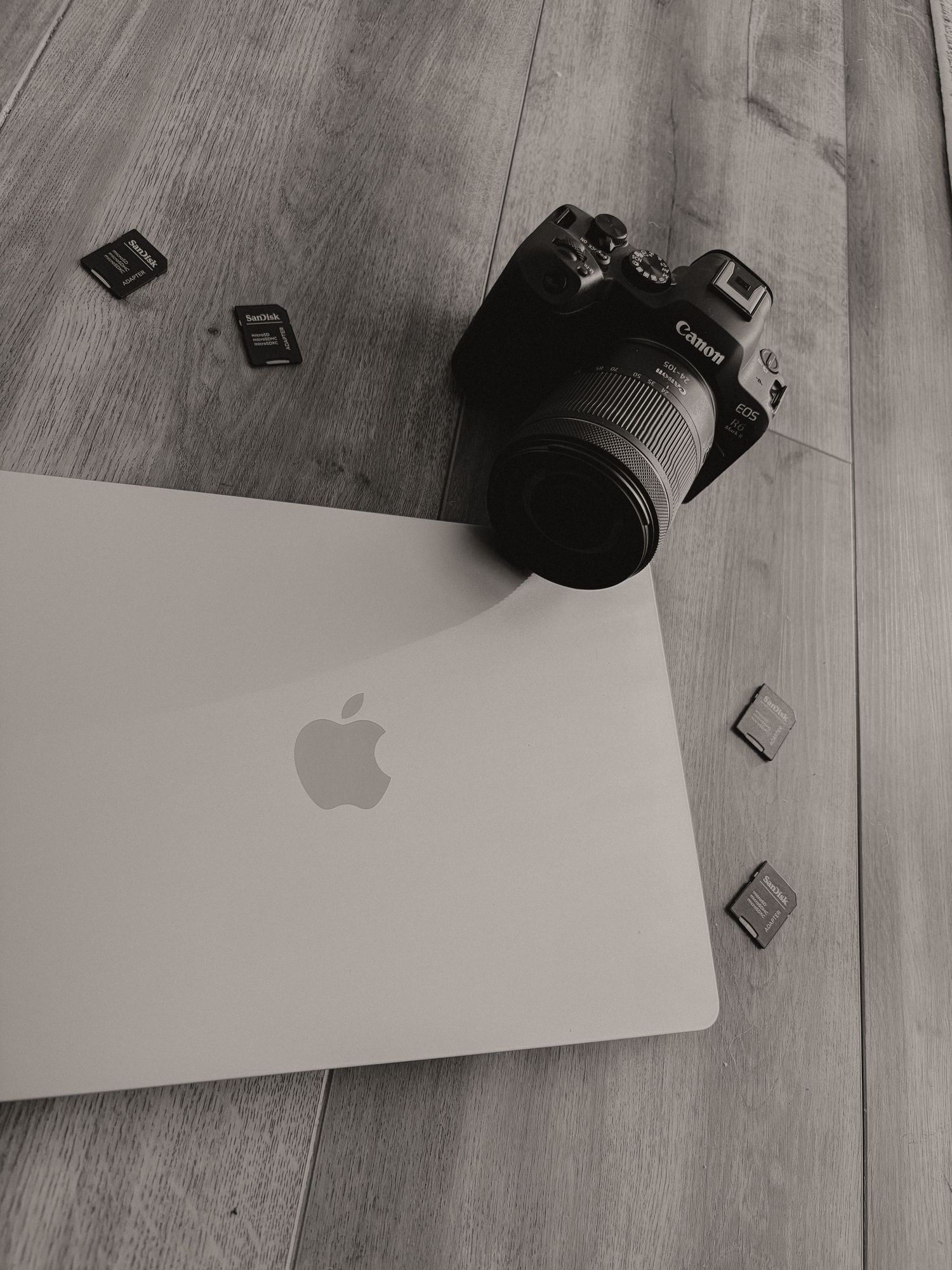 Black and white flat lay of Canon DSLR camera, SD cards, and Apple MacBook on wooden floor.