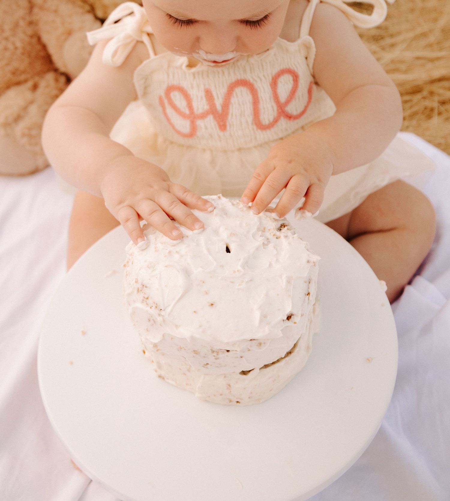 Baby girl in 'one' romper smashing white birthday cake with both hands during first birthday cake smash.