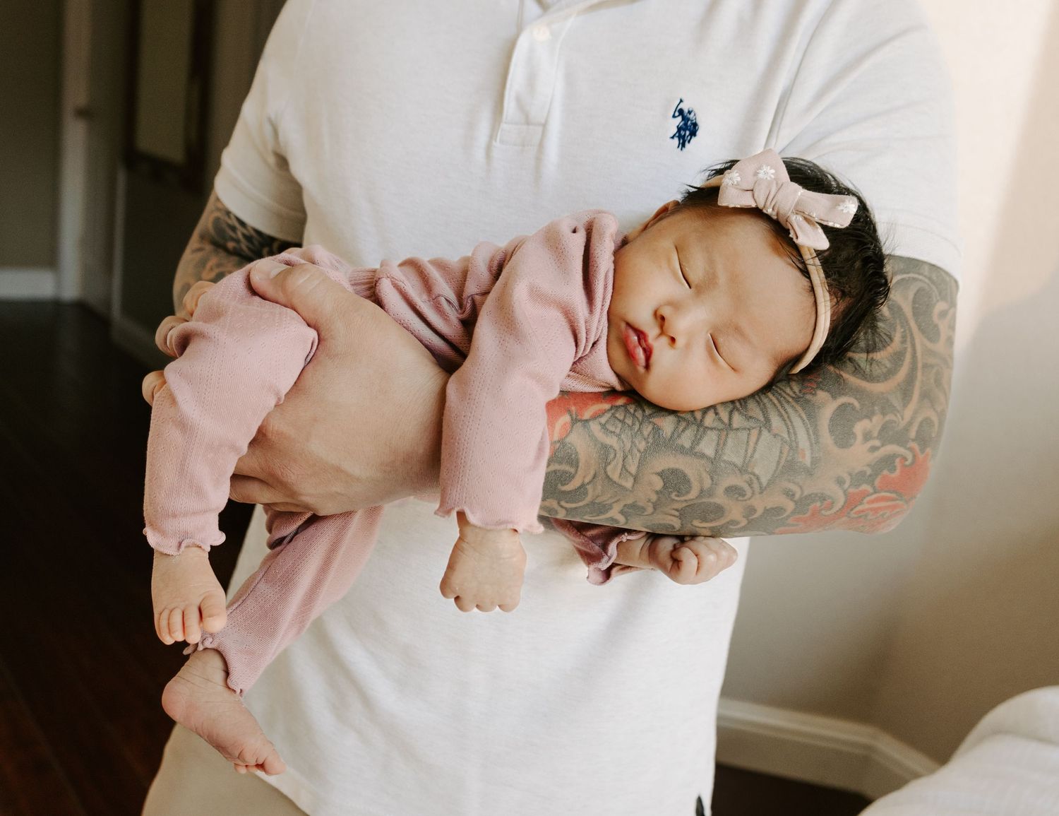 Tattooed father in white polo shirt cradling sleeping newborn baby girl in pink.