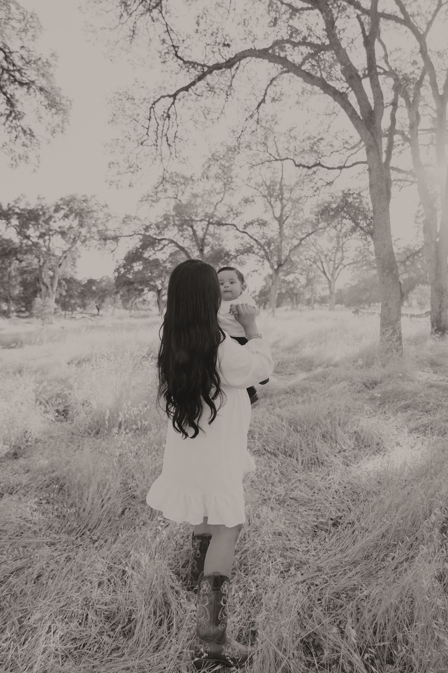 A mother with long dark hair in a white dress holds her baby outdoors among golden grass and bare trees.