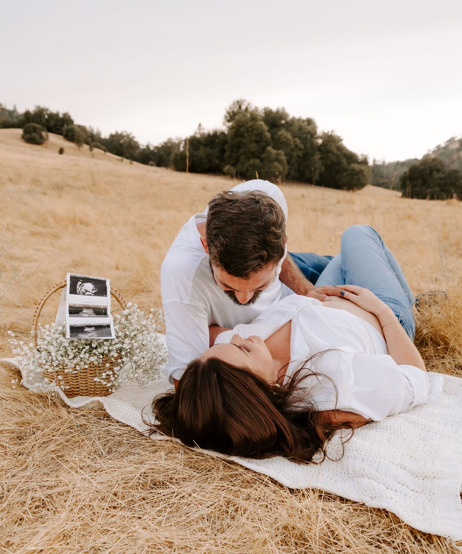 Couple sharing a romantic moment on a blanket in a golden field with baby sonogram photos nearby.