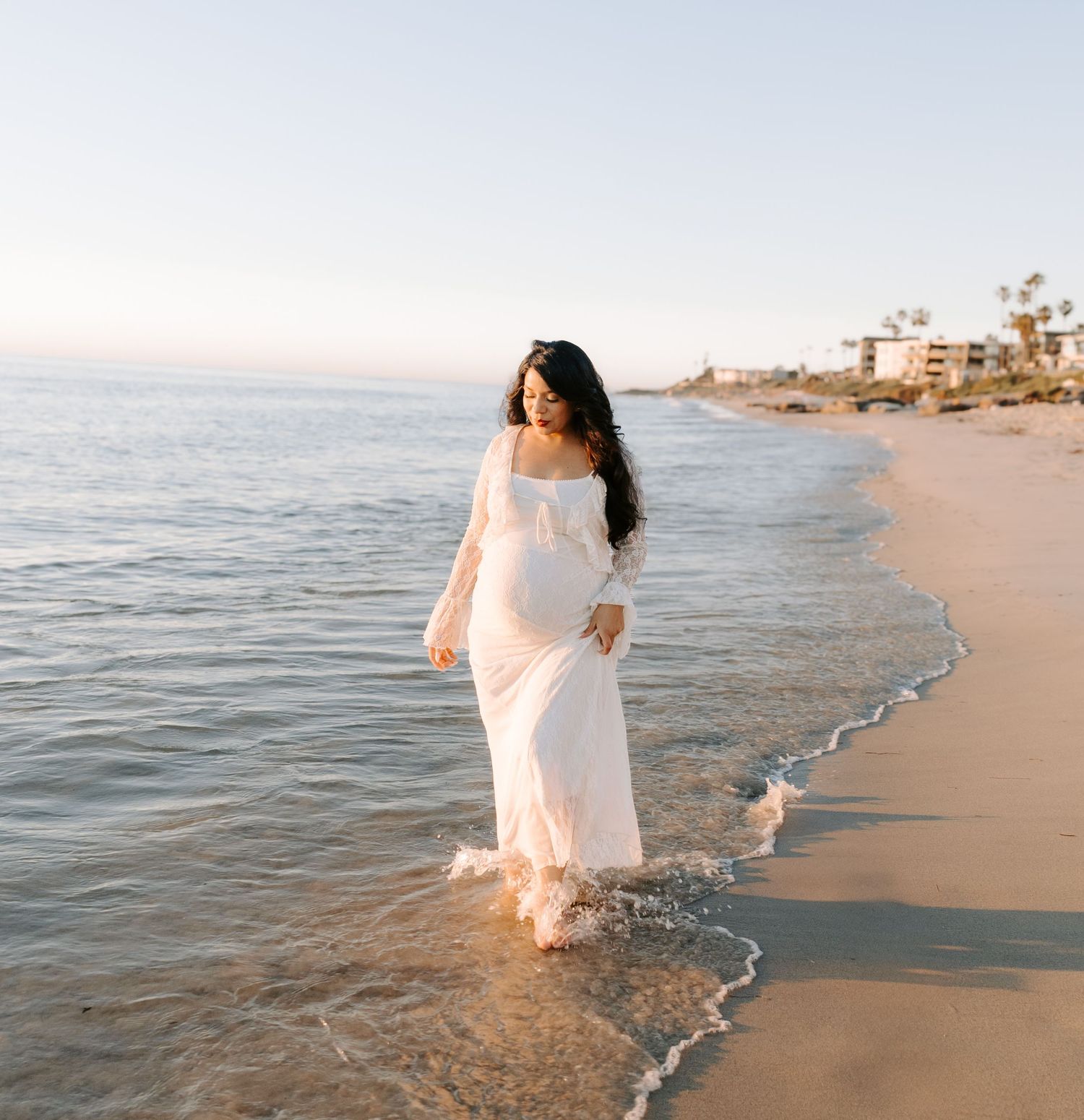 Pregnant woman in flowing white off-shoulder gown walks gracefully along ocean shoreline at sunset.