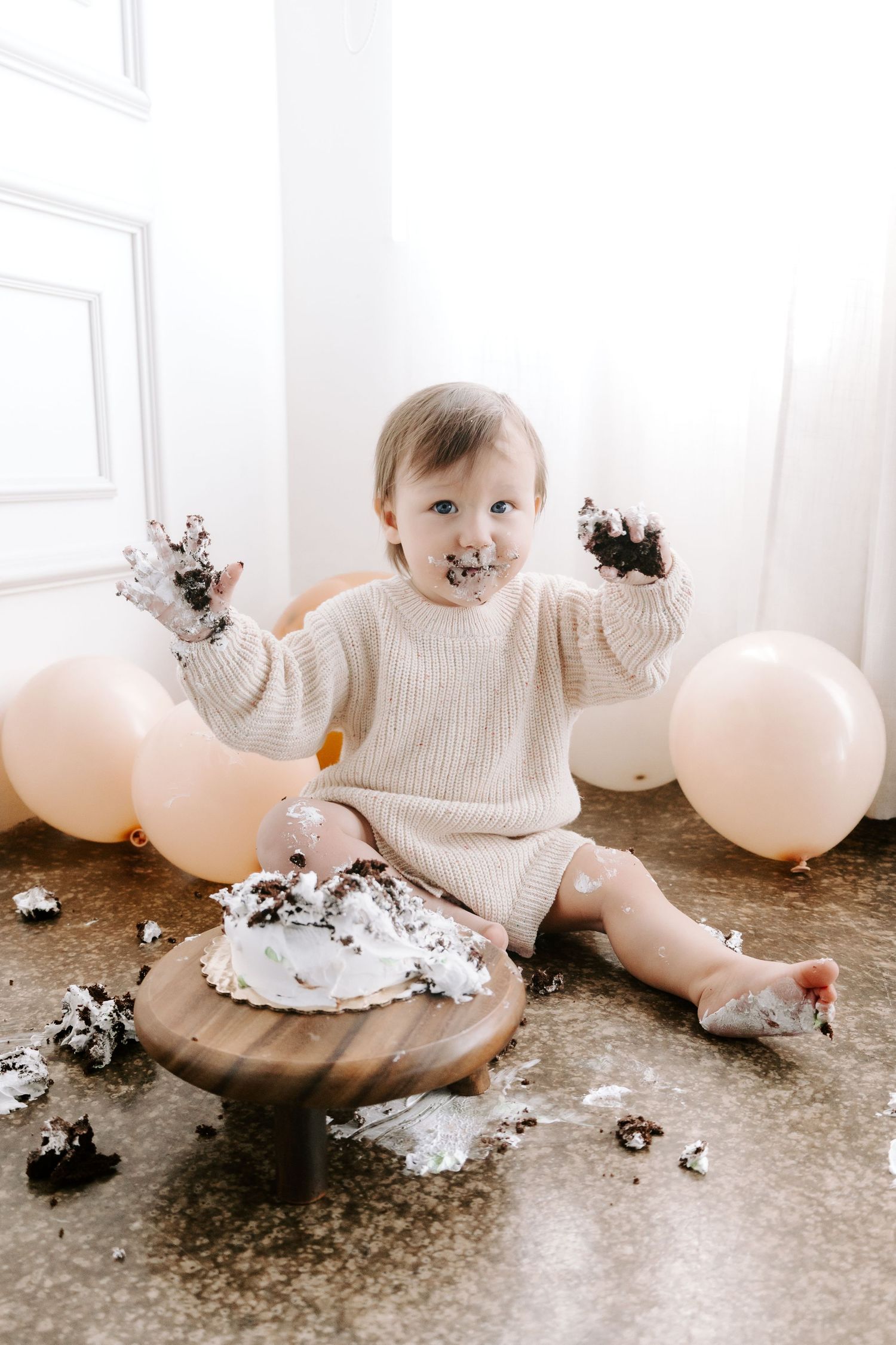 Joyful baby girl in beige sweater smashes first birthday cake surrounded by blush balloons.